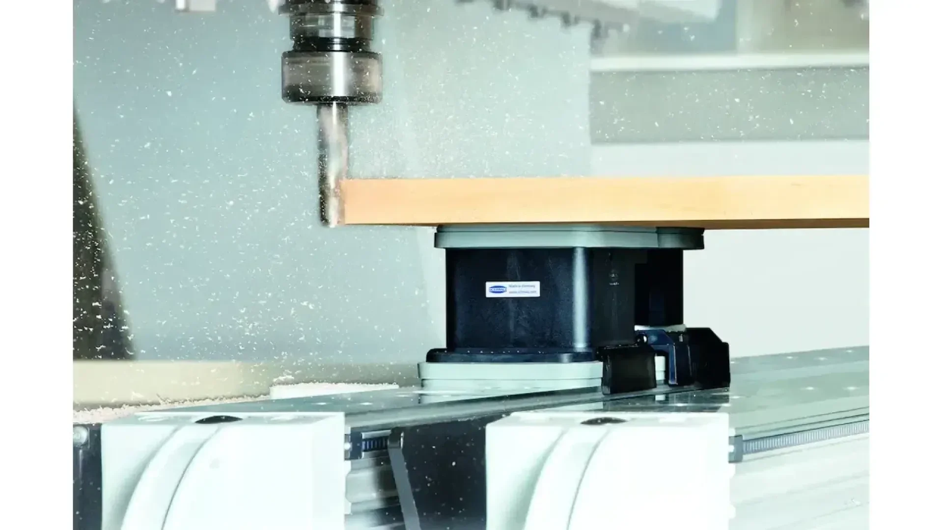 Une machine à commande numérique perce une pièce de bois, avec des particules de sciure qui volent autour ; le bois est fixé sur un support à vide sous le foret dans un environnement industriel.