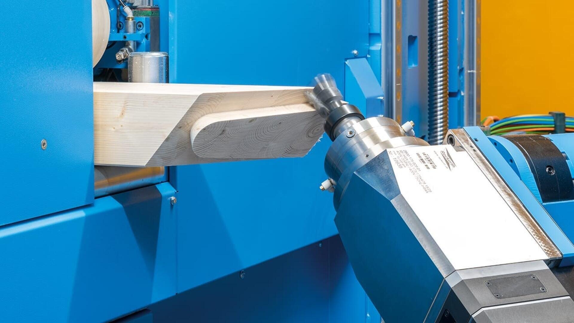 Gros plan d'une machine industrielle à commande numérique qui coupe et façonne un morceau de bois clair. Les composants bleus de la machine et le fond jaune sont visibles.