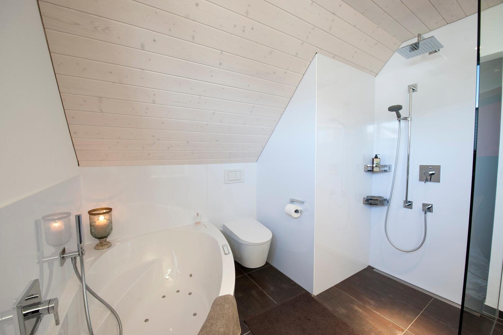 Modernes Badezimmer mit einer schrägen Holzdecke, weißen Wänden, einer runden Badewanne, einer Wandtoilette, Kerzen und einer begehbaren Dusche mit verchromten Armaturen und Regalen für Toilettenartikel.