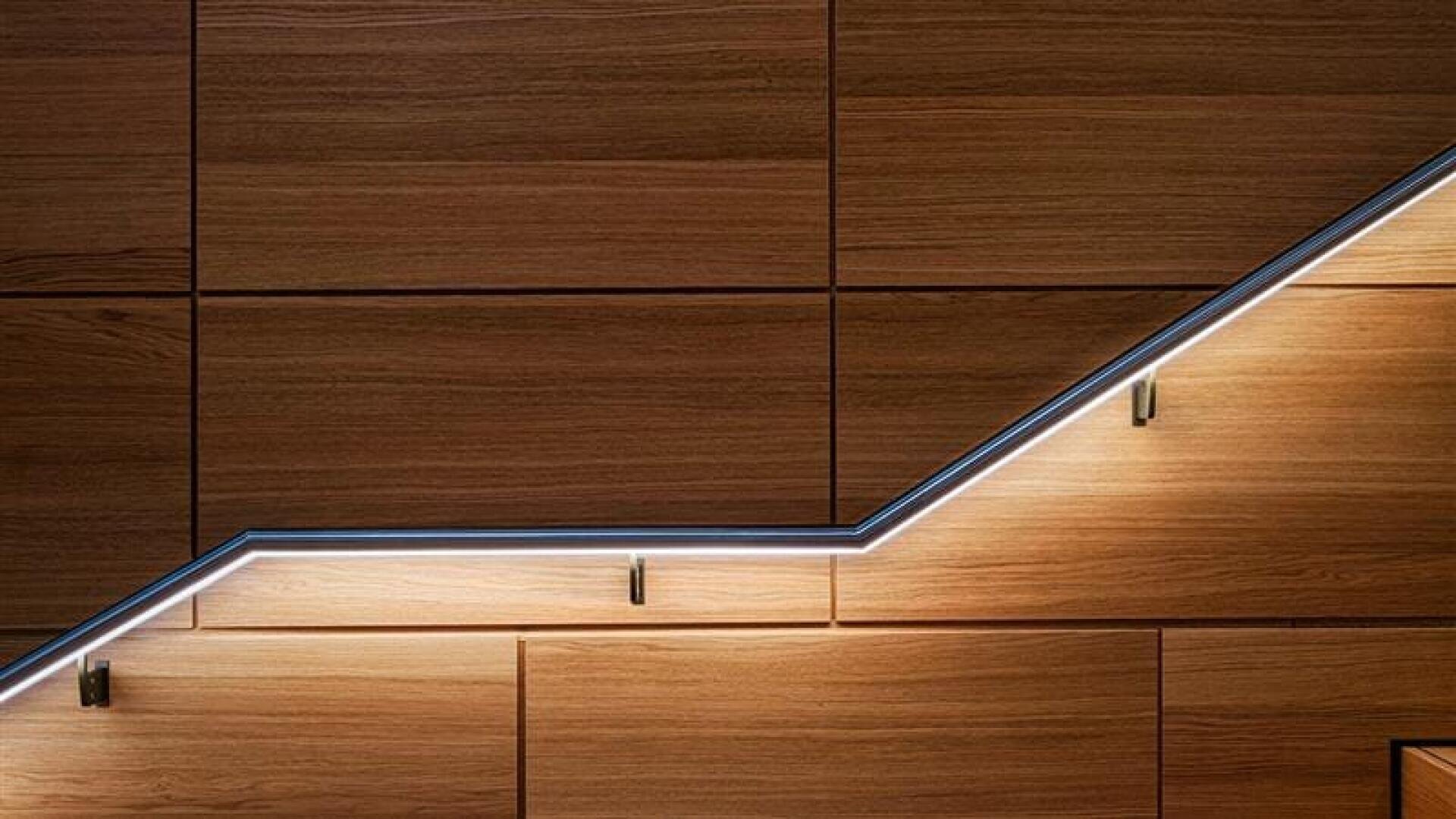 Un mur moderne en bois avec des panneaux rectangulaires, doté d'une main courante élégante et diagonale éclairée par une bande de lumière LED et soutenue par de petits supports métalliques.