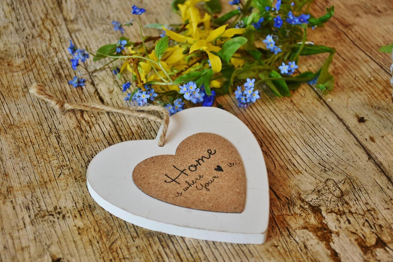 Ein weißes Holzherz mit einem Seil und dem Schriftzug "Home is where your heart is" ruht auf einer rustikalen Holzfläche, neben einem kleinen Strauß gelber und blauer Blumen.