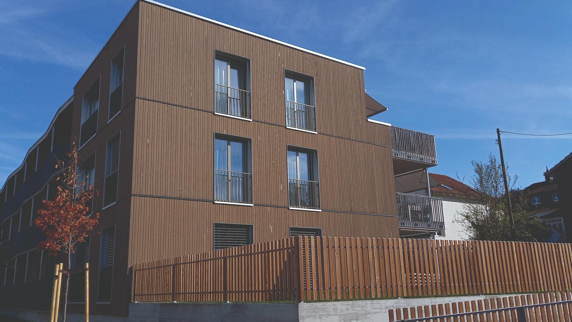 Ein modernes dreistöckiges Wohnhaus mit Holzvertäfelung, großen Fenstern und kleinen Balkonen, umgeben von einem passenden Holzzaun unter einem klaren blauen Himmel.