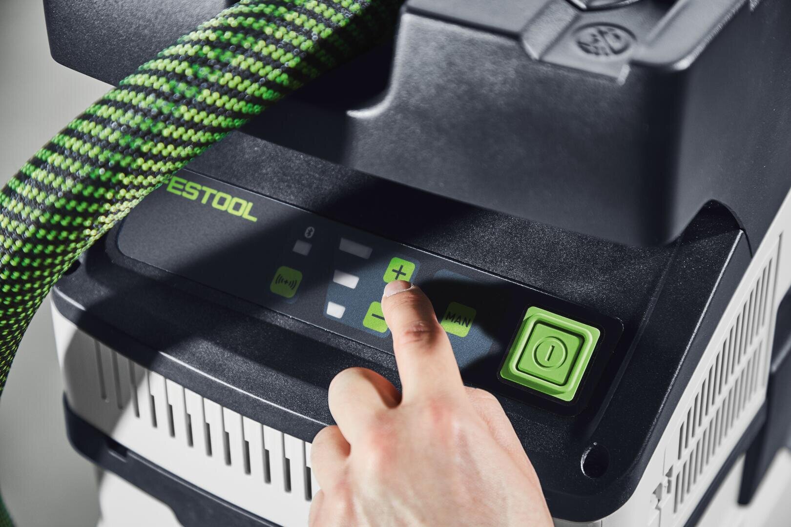 Eine Hand drückt die grüne Plustaste auf dem Bedienfeld eines Festool Staubsaugers oder Staubabscheiders. Auf dem Bedienfeld sind LED-Anzeigen und grüne Akzente zu sehen, und neben den Bedienelementen ist eine grüne Einschalttaste zu sehen.