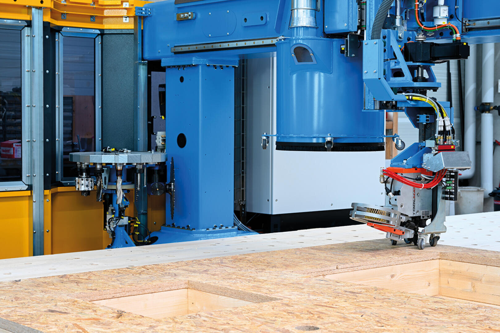 Eine große blaue CNC-Industriemaschine schneidet rechteckige Öffnungen in eine Holzplatte, mit sichtbaren Metallteilen und Verkabelung in einer Fabrikumgebung.
