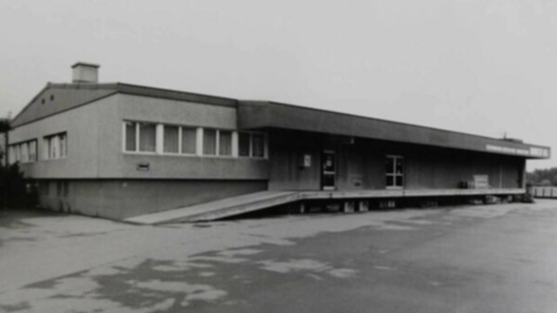 Ein Schwarz-Weiß-Foto eines niedrigen, rechteckigen Betongebäudes mit mehreren Fenstern und einer langen Laderampe an der Vorderseite. Die Struktur ist schlicht und industriell, mit einer leeren gepflasterten Fläche im Vordergrund.