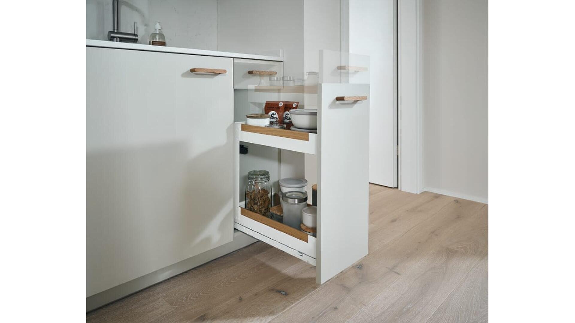 Une cuisine moderne avec un meuble coulissant blanc ouvert, présentant deux étagères contenant des bocaux, des boîtes de conserve et des bols. La cuisine est dotée d'un sol en bois clair et d'un design minimaliste.
