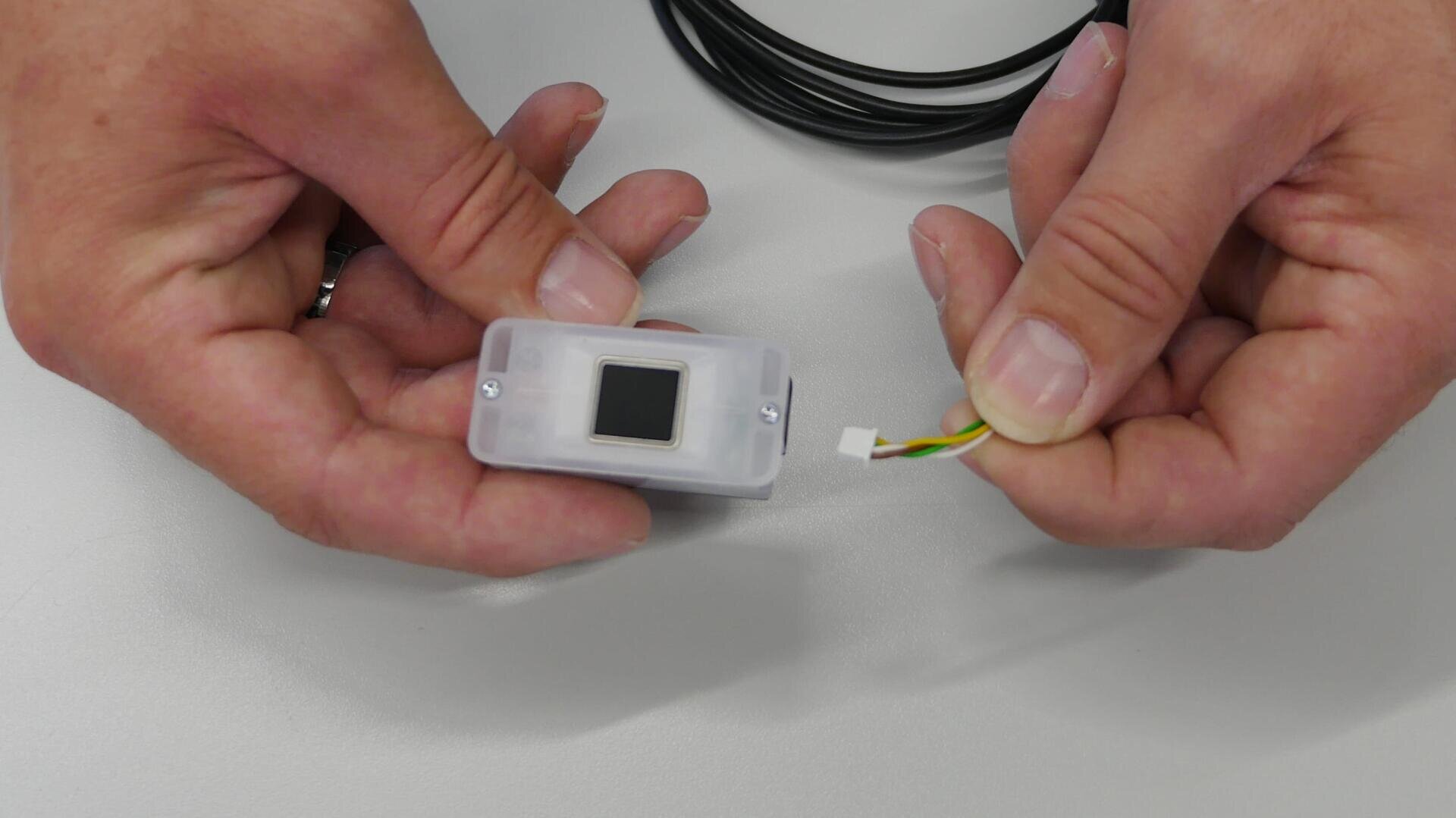 Eine Person hält ein kleines elektronisches Gerät mit einem schwarzen quadratischen Sensor in der einen Hand und ein Kabel mit einem weißen Stecker und farbigen Drähten in der anderen Hand.