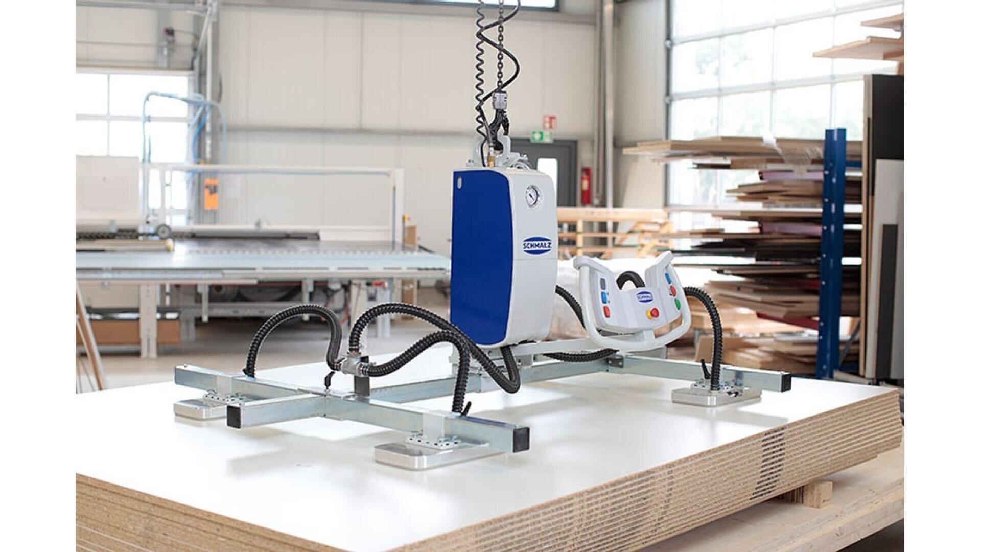 Une machine de levage par le vide munie de ventouses soulève de grandes planches de bois dans un atelier spacieux et bien éclairé, avec des planches empilées et des équipements de travail du bois à l'arrière-plan.