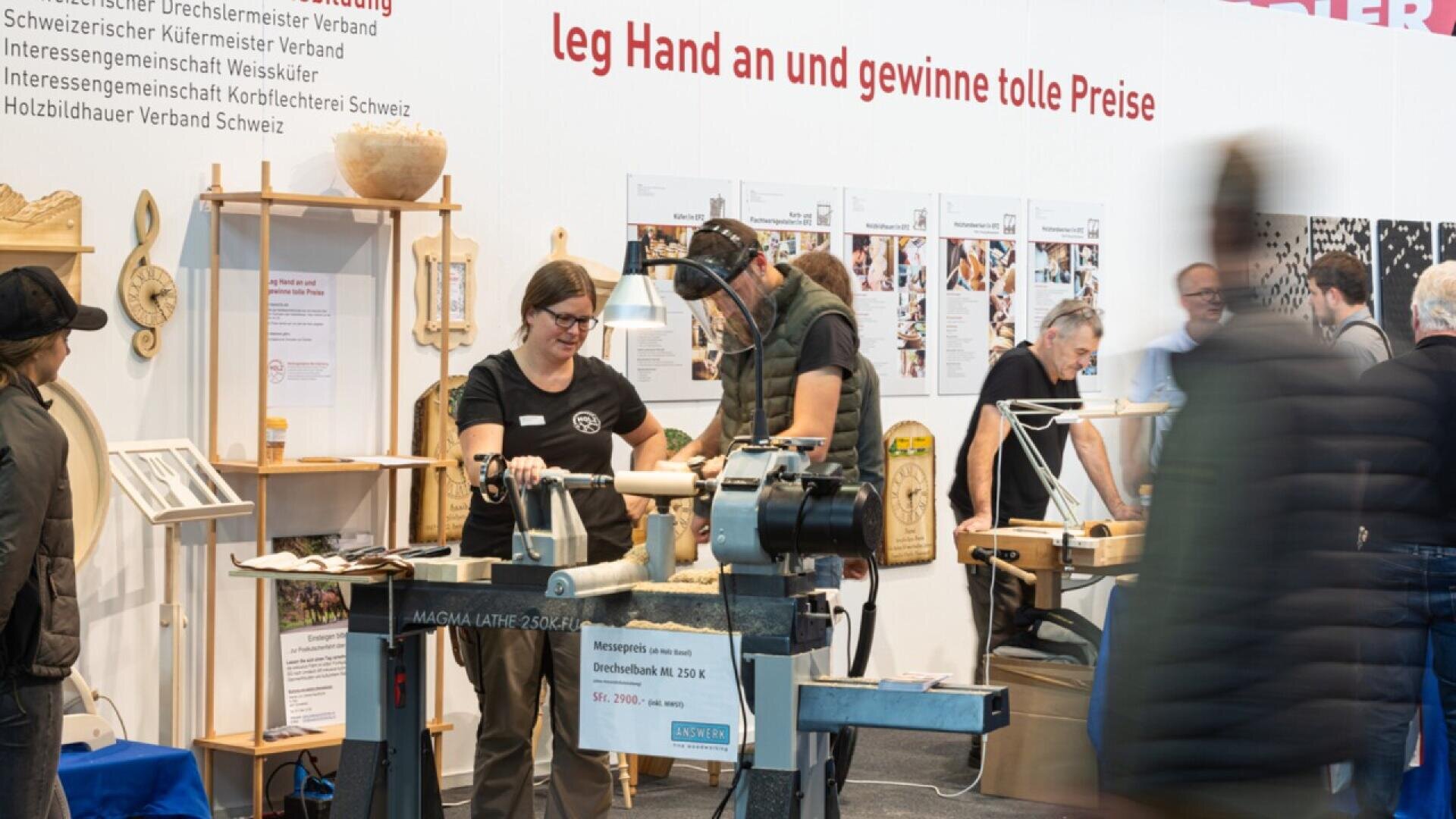 Des personnes participant à une démonstration de travail du bois utilisent un tour, tandis que d'autres observent à l'arrière-plan. Des objets et des outils en bois sont exposés autour d'eux. Une bannière au-dessus offre une chance de gagner des prix.