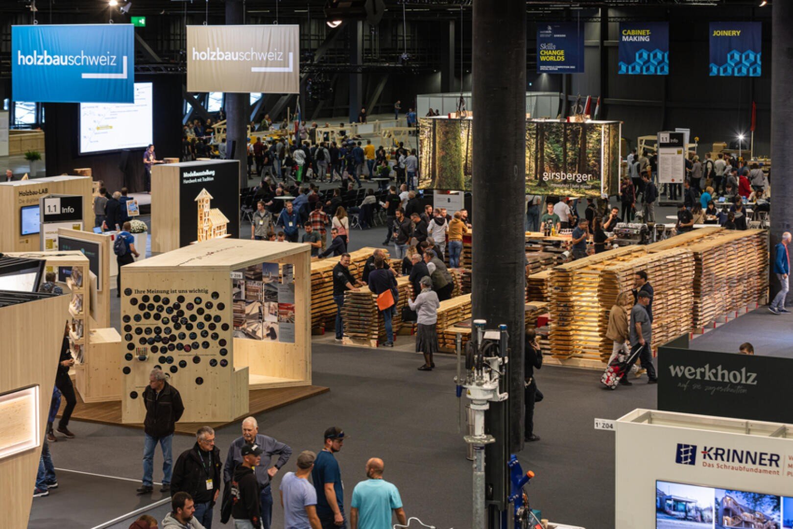 In einer belebten Ausstellungshalle gibt es Holzkonstruktionen, Informationsstände und Ausstellungen zum Thema Holzbau. Viele Menschen gehen umher, erkunden die Exponate und unterhalten sich mit den Referenten. Große Schilder hängen über den verschiedenen Ständen.