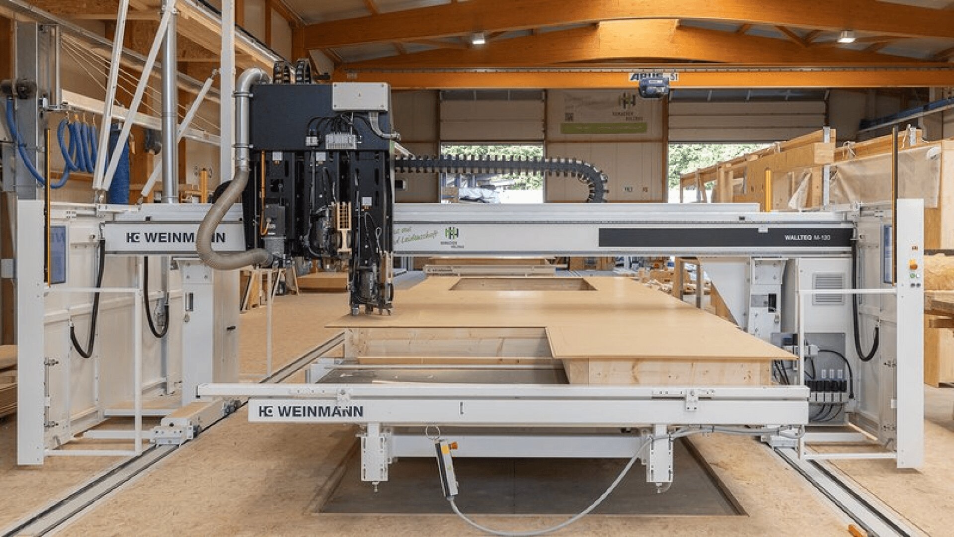 Eine große, automatisierte CNC-Holzbearbeitungsmaschine steht in einer geräumigen, modernen Werkstatt, in der im Hintergrund Holzbalken und verschiedene Holzbearbeitungsmaterialien und -geräte zu sehen sind.