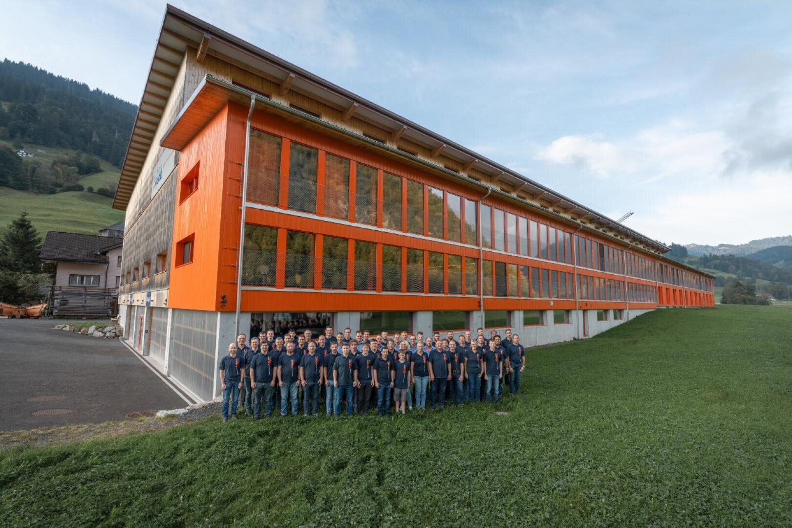Un grand groupe de personnes portant des chemises assorties se tient dans l'herbe devant un long bâtiment moderne aux murs extérieurs orangés et vitrés, dans un paysage pittoresque et vallonné.