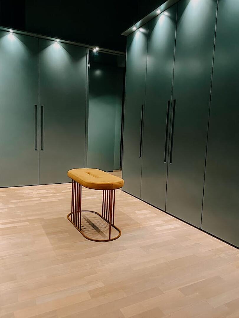 Ein moderner begehbarer Kleiderschrank mit dunkelgrünen Schränken, eleganten schwarzen Griffen, eingebauter Deckenbeleuchtung und einer braunen gepolsterten Bank mit Metallrahmen auf hellem Holzfußboden.
