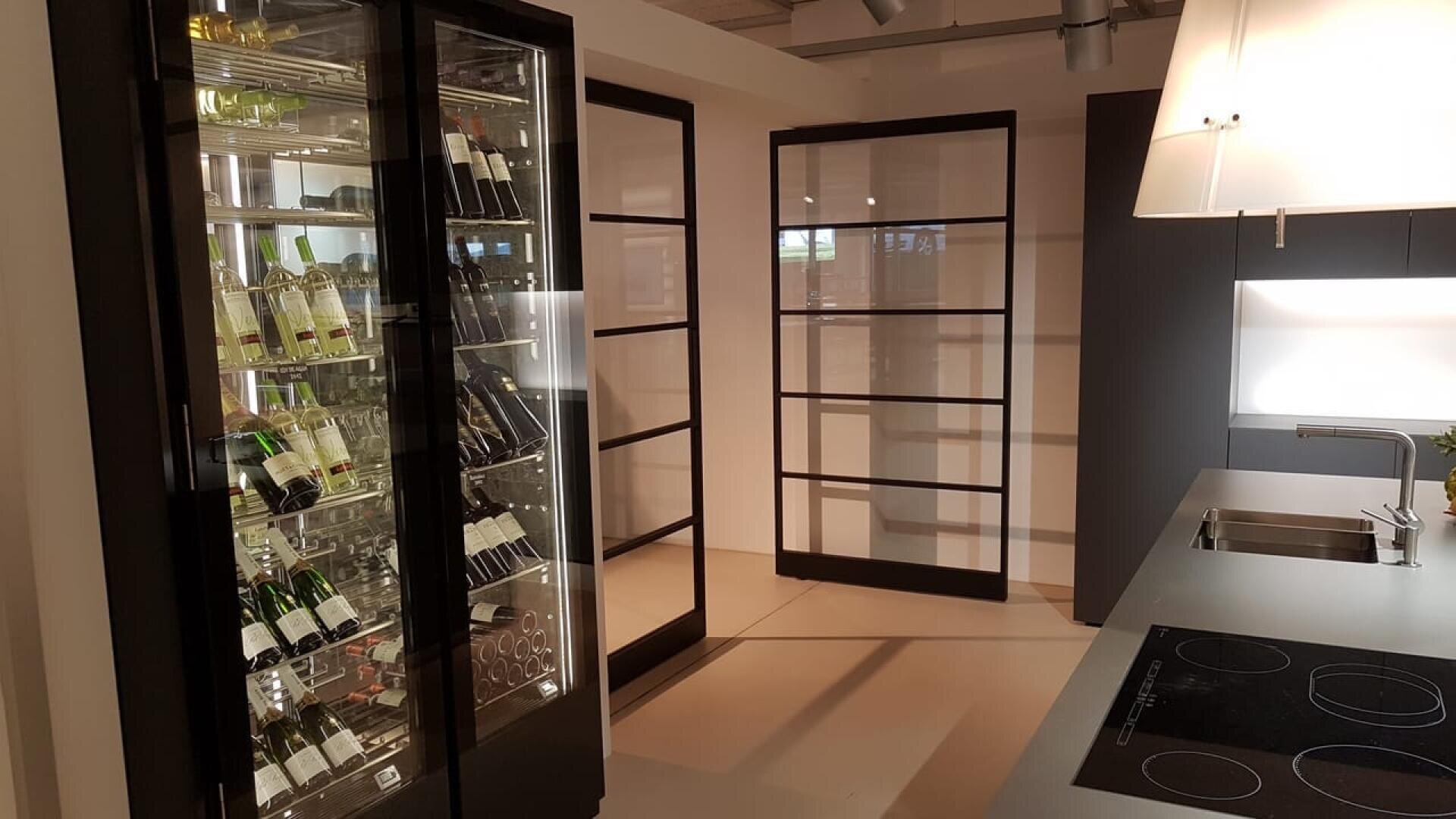 La cuisine moderne est équipée d'un réfrigérateur à vin vitré rempli de bouteilles, d'armoires noires élégantes, d'une cuisinière électrique et d'un évier en acier inoxydable, le tout dans un décor et un éclairage minimalistes.