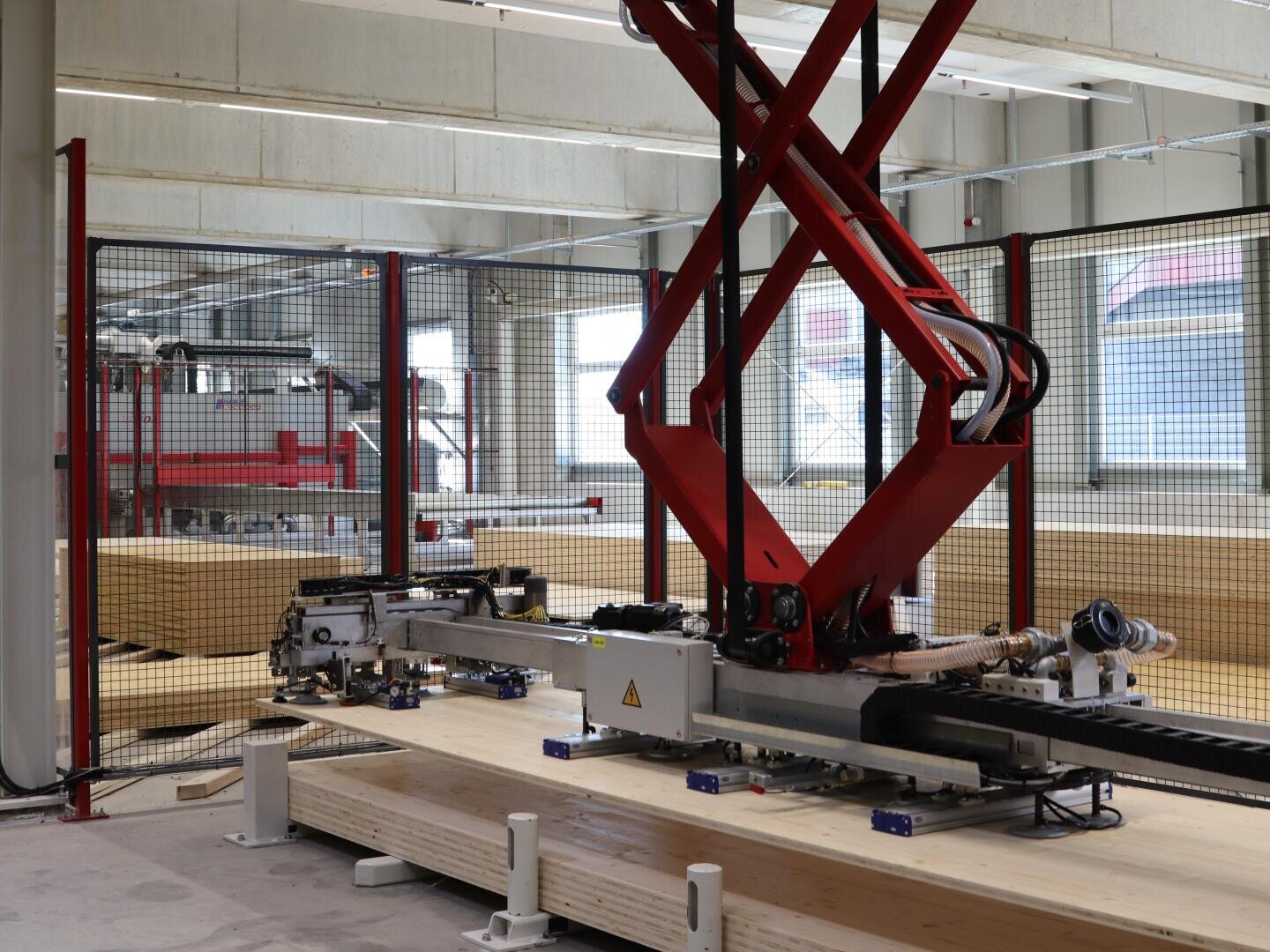 Un bras robotisé avec des supports rouges fonctionne dans une installation industrielle moderne, manipulant de grandes planches de bois derrière une barrière de sécurité avec des machines et des fenêtres en arrière-plan.