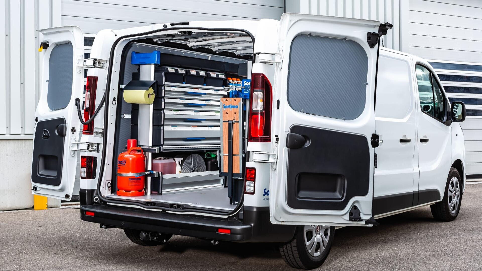 Ein weißer Kleintransporter mit geöffneten Hecktüren, die den Blick auf geordnete Regale, Schubladen, Werkzeuge, eine rote Gasflasche und verschiedene Ausrüstungsgegenstände freigeben, die im Inneren zu Arbeits- oder Reparaturzwecken gelagert werden.