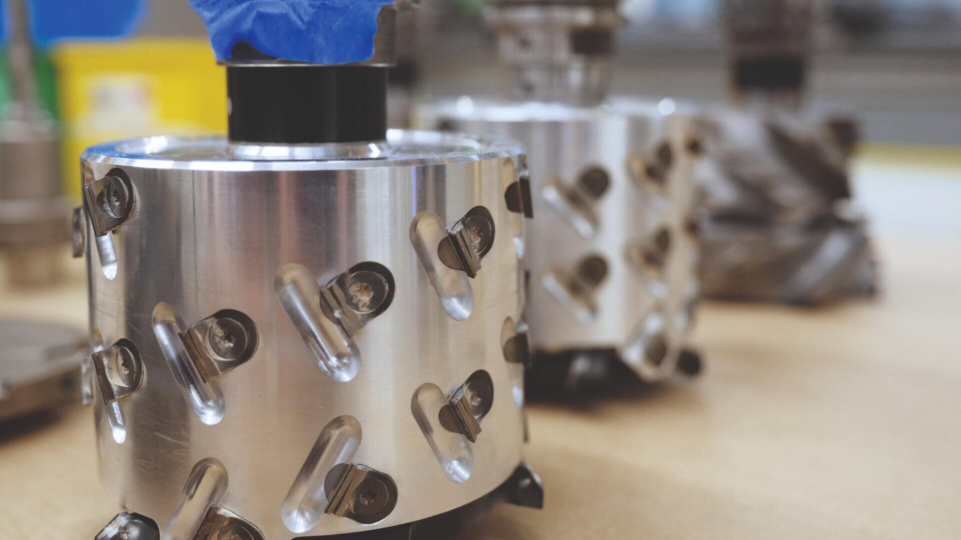 Nahaufnahme eines glänzenden zylindrischen Industriewerkzeugs aus Metall mit scharfkantigen Einsätzen und Bolzen, das in einer Werkstatt auf eine Holzfläche gestellt wird, wobei ein anderes ähnliches Werkzeug im Hintergrund verschwimmt.