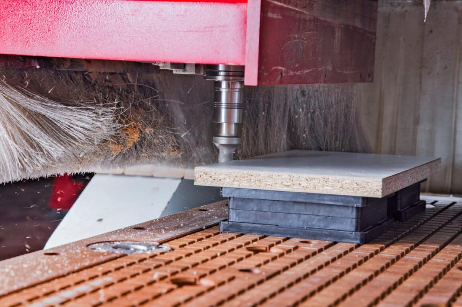 Eine CNC-Fräsmaschine schnitzt oder schneidet ein rechteckiges Stück Spanplatte, das auf einem Arbeitstisch mit einem Gittermuster befestigt ist. Das Schneidewerkzeug befindet sich direkt über der Platte und ist betriebsbereit.