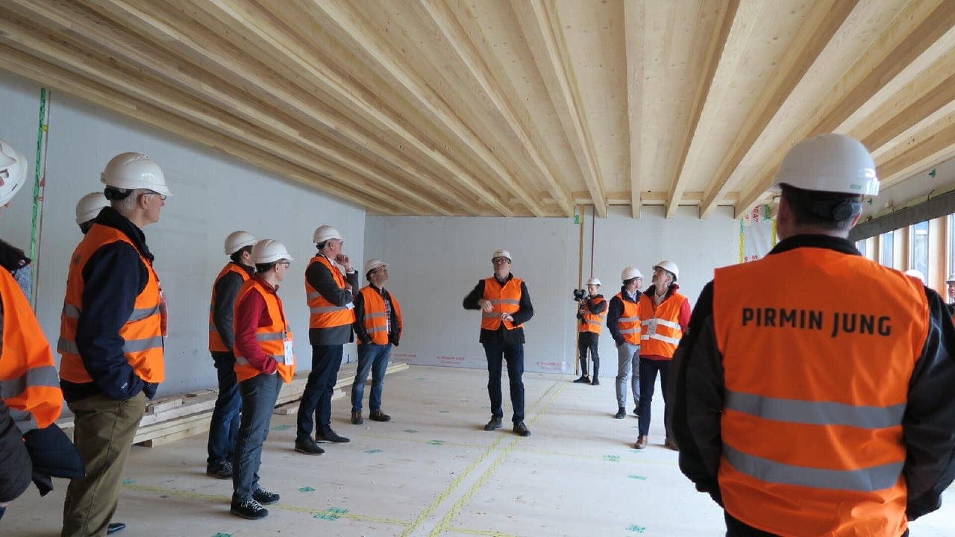 Un groupe de personnes portant des gilets de sécurité orange et des casques blancs se tient à l'intérieur d'un bâtiment en construction, écoutant un orateur. L'un des gilets porte l'inscription PIRMIN JUNG au dos. L'espace a des poutres en bois apparentes.