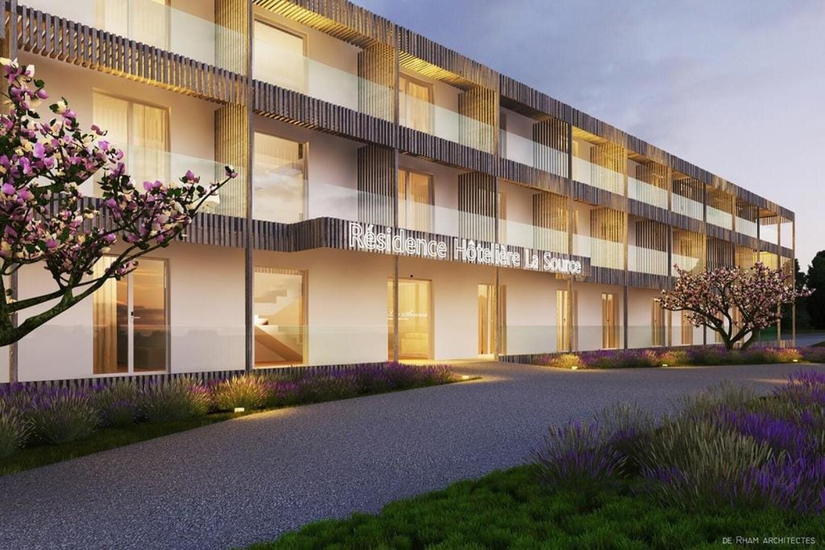 Ein modernes, zweistöckiges Gebäude mit Holzakzenten, großen Glasbalkonen, beleuchteten Eingängen und blühenden Bäumen entlang eines Weges vor dem Gebäude.