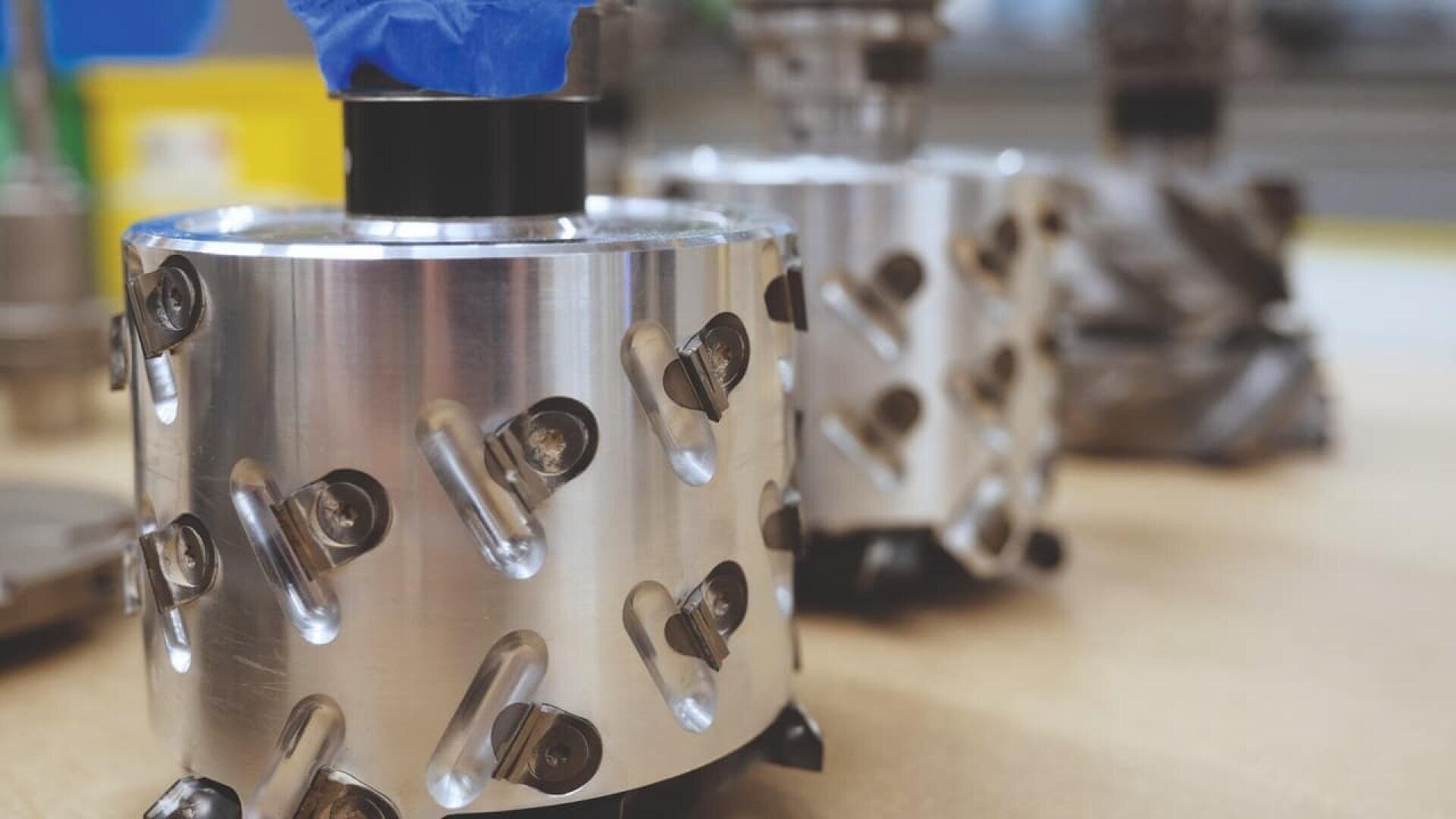 Gros plan sur un outil métallique cylindrique et brillant, dont la surface est recouverte de multiples vis inclinées, posé sur un établi. Un autre outil similaire est flou à l'arrière-plan.