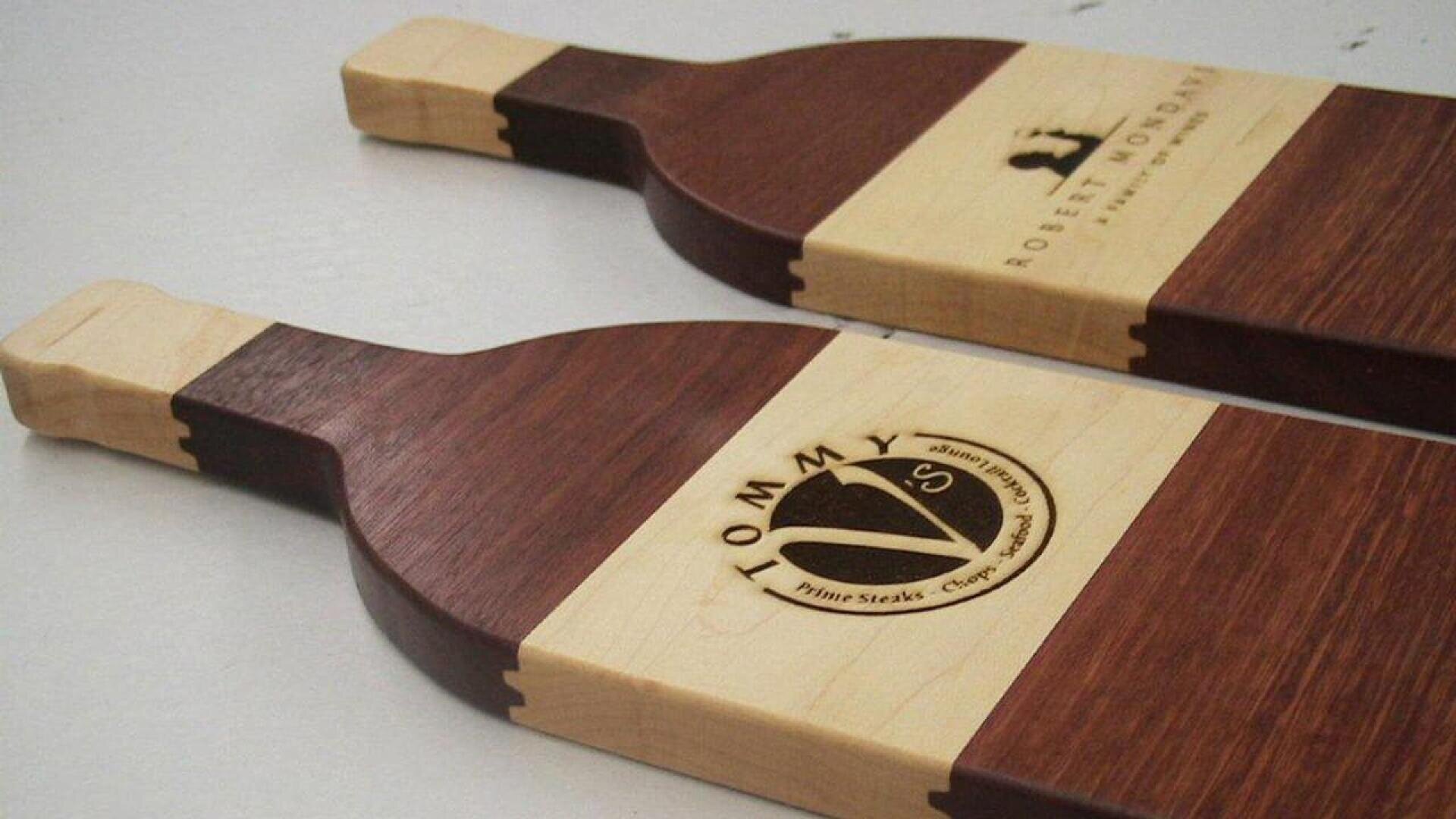 Deux planches à découper en bois avec des motifs contrastés en bois clair et foncé, des poignées rectangulaires et des logos gravés près de la partie supérieure. Les gravures comprennent du texte et des éléments graphiques simples.