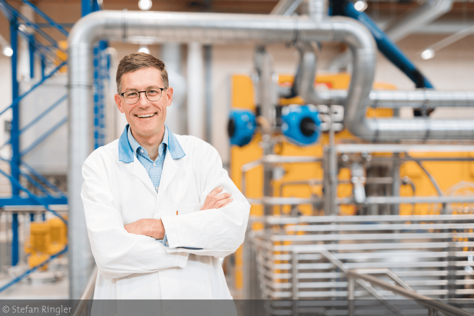 Ein Mann mit Brille und weißem Laborkittel steht lächelnd mit verschränkten Armen in einer Industrieanlage mit Metallrohren und gelben Maschinen im Hintergrund.