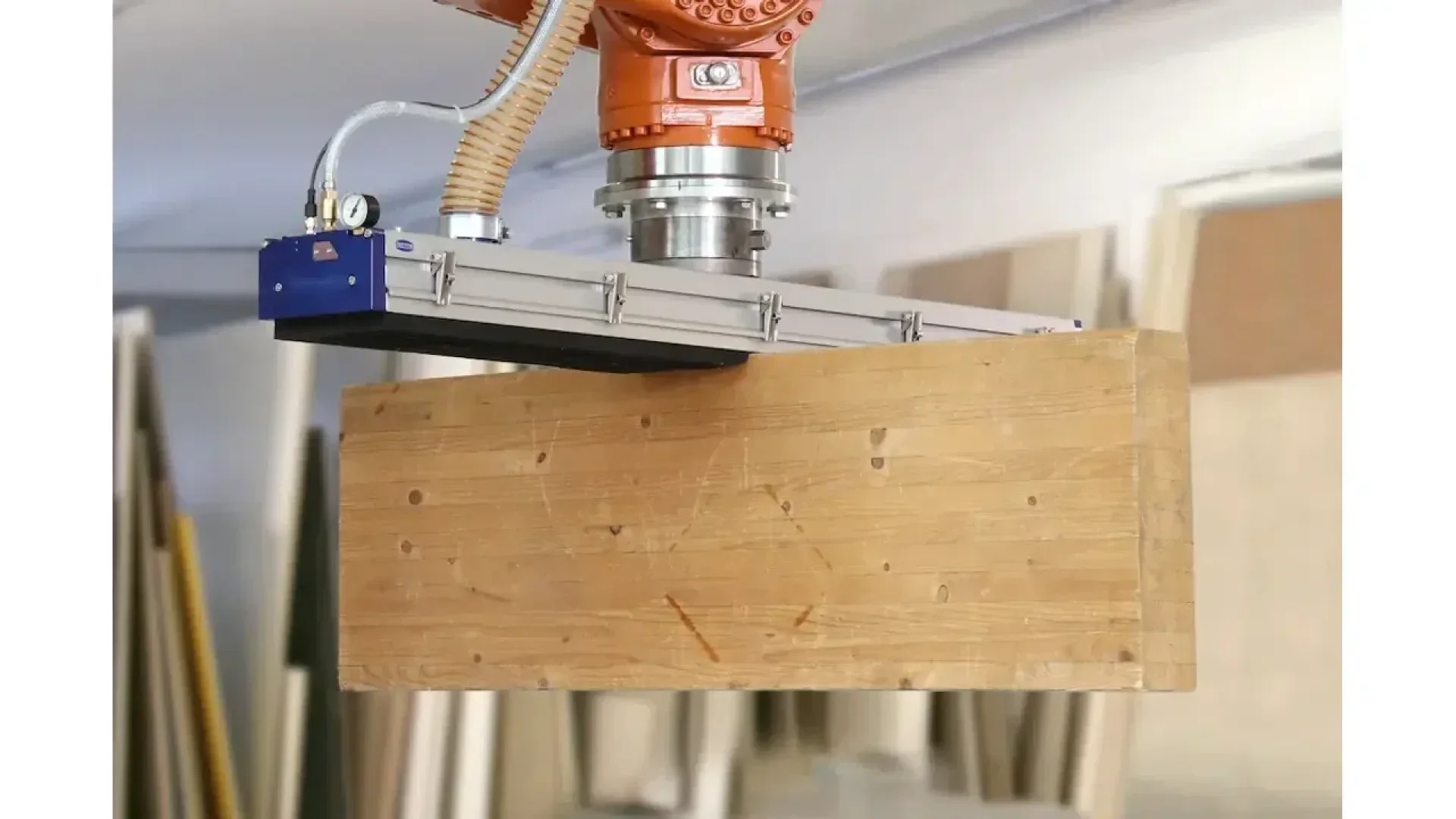 Un bras robotisé équipé d'un dispositif d'aspiration soulève une grande planche de bois rectangulaire dans un environnement industriel ou un atelier en intérieur.