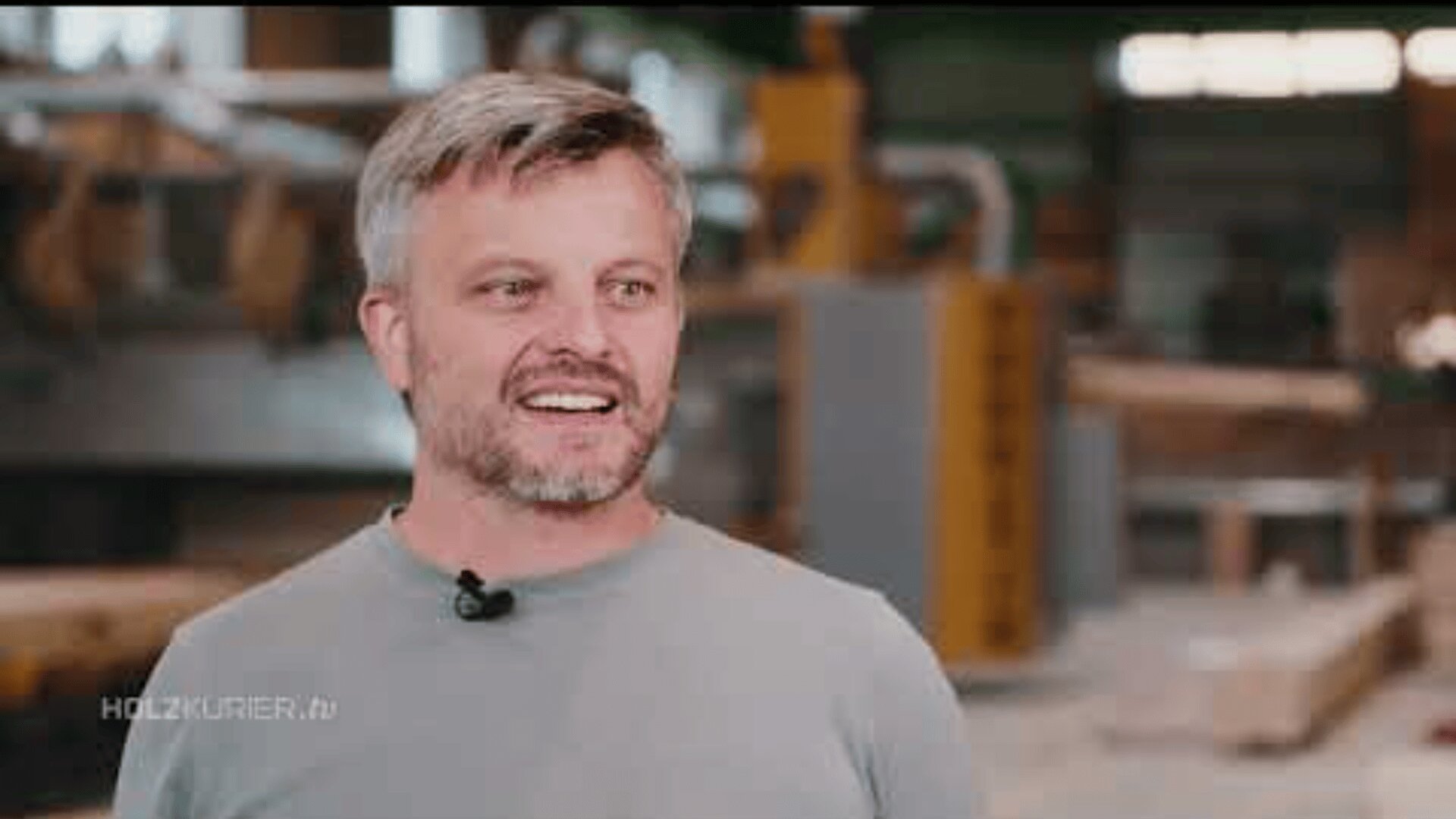 Ein Mann mit kurzen grauen Haaren und Bart, der ein hellgraues Hemd und ein Mikrofon trägt, lächelt in einer Werkstatt mit unscharfen Industrieanlagen im Hintergrund. In der unteren linken Ecke erscheint der Schriftzug HOLZKURIER.tv.