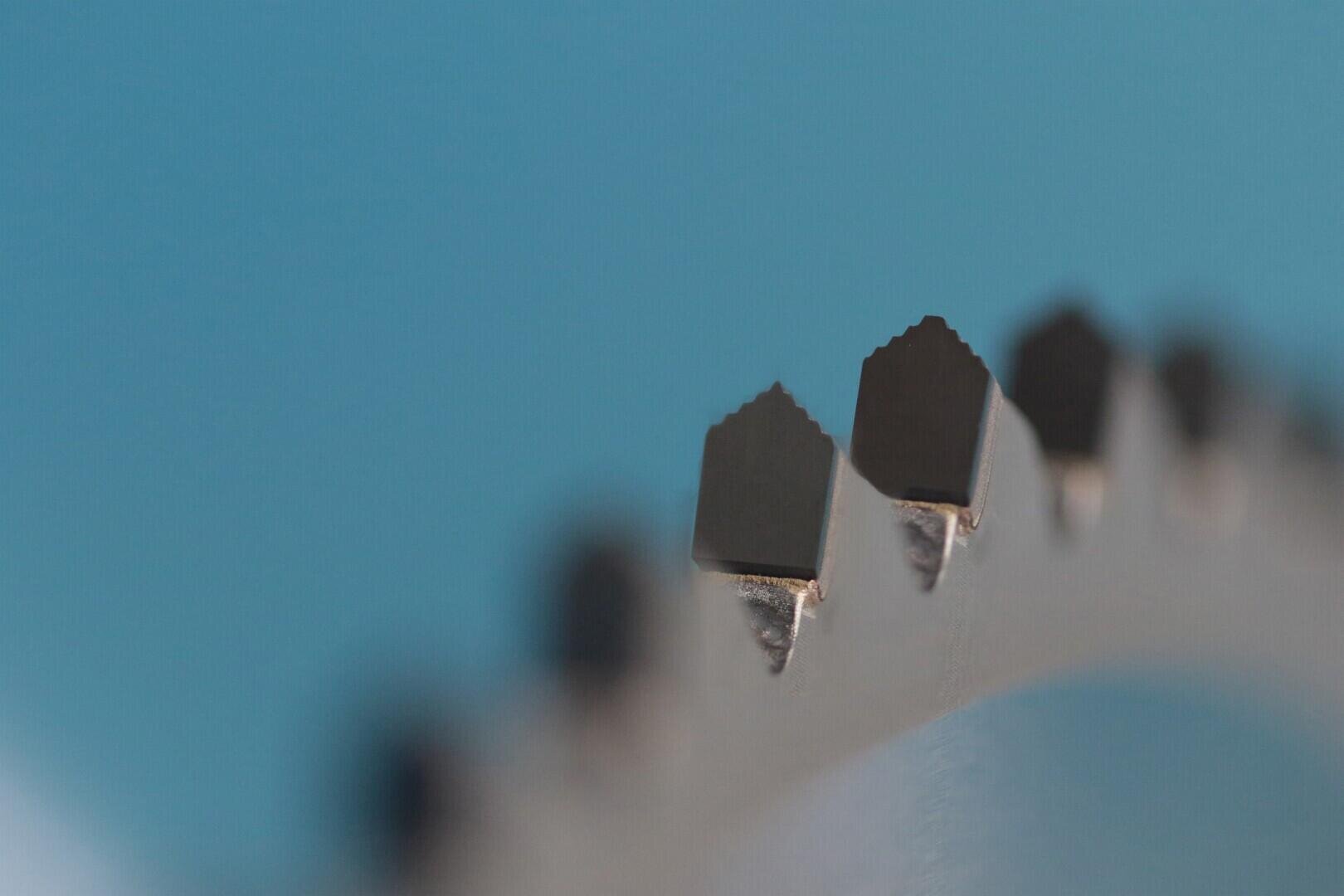 Nahaufnahme der gezackten, spitzen Kante eines Metallzahnrads oder Sägeblatts vor einem glatten blauen Hintergrund, die feine Details der Metallzähne zeigt.