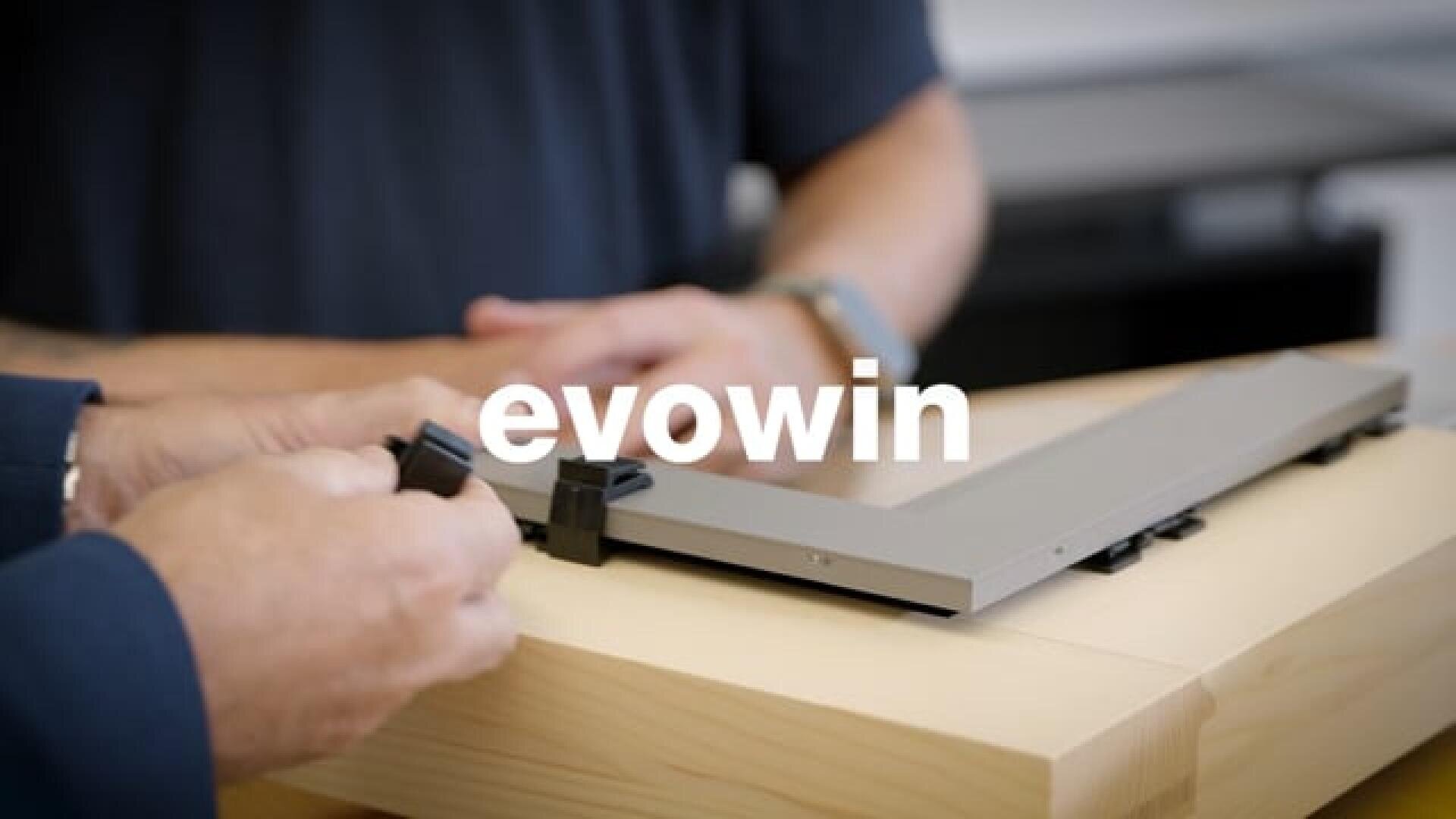 Zwei Personen montieren einen rechteckigen Metallrahmen mit schwarzen Klammern auf einem Holztisch, wobei das Wort evowin in weißer Schrift in der Mitte des Bildes eingeblendet ist.