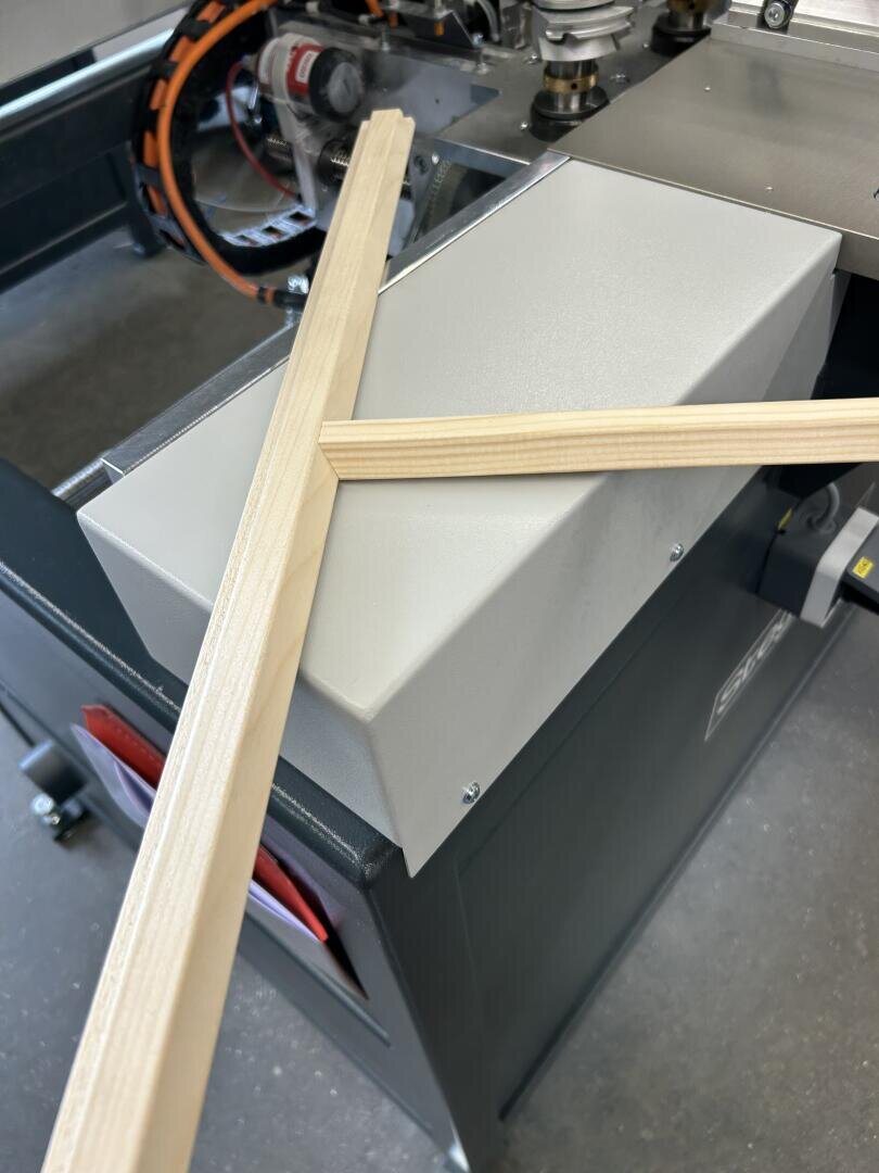Zwei Holzleisten mit auf Gehrung geschnittenen Enden sind in einer X-Form auf einer industriellen Holzbearbeitungsmaschine aus Metall angeordnet. Die Maschine verfügt über Knöpfe und sichtbare mechanische Komponenten im Hintergrund.
