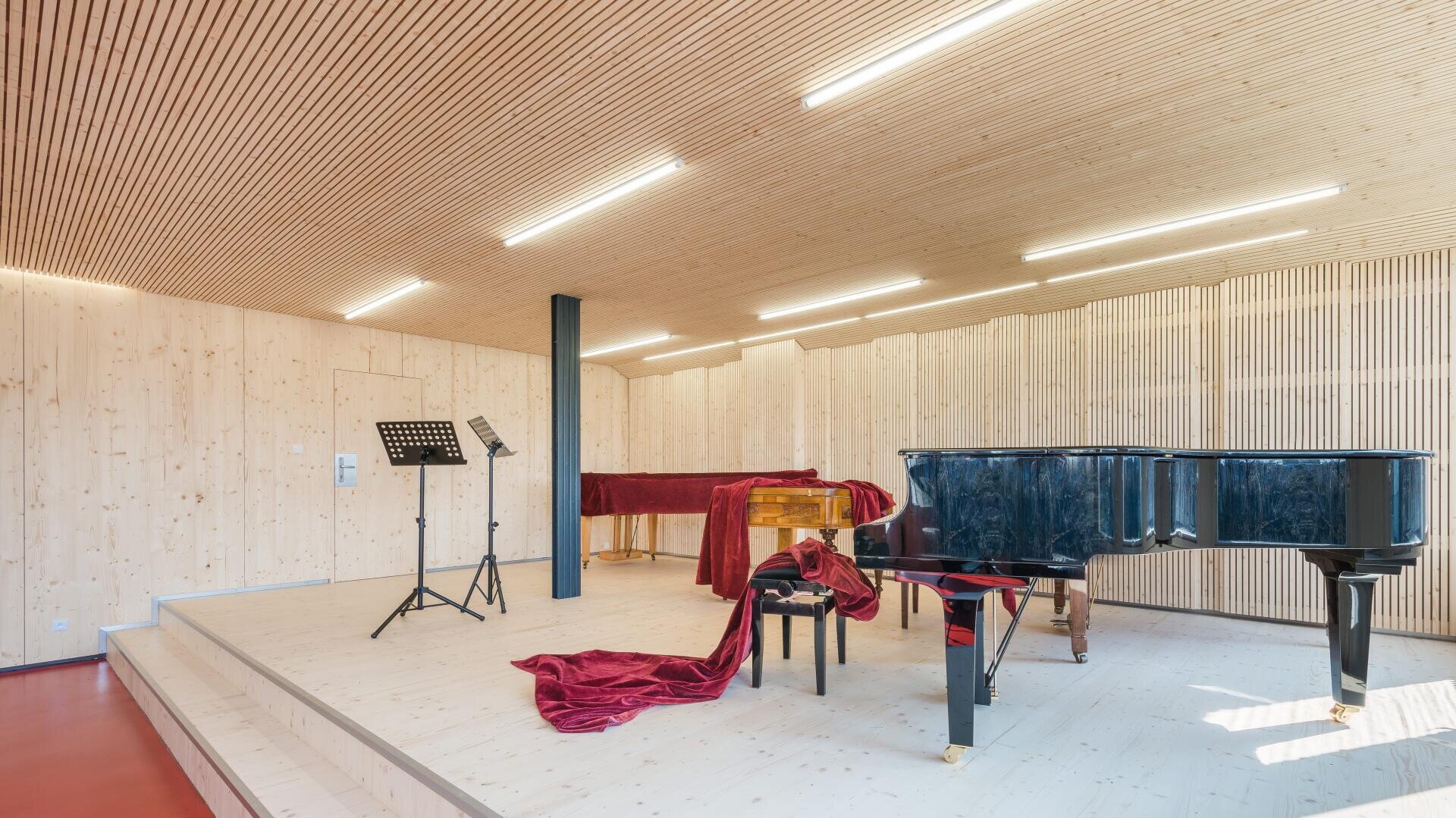 Ein modernes Musikzimmer mit Holzwänden und -decke, in dem ein schwarzer Flügel, ein Cembalo, Notenständer und roter Samtstoff über den Instrumenten drapiert sind. Helle Deckenleuchten erhellen die Bühne.