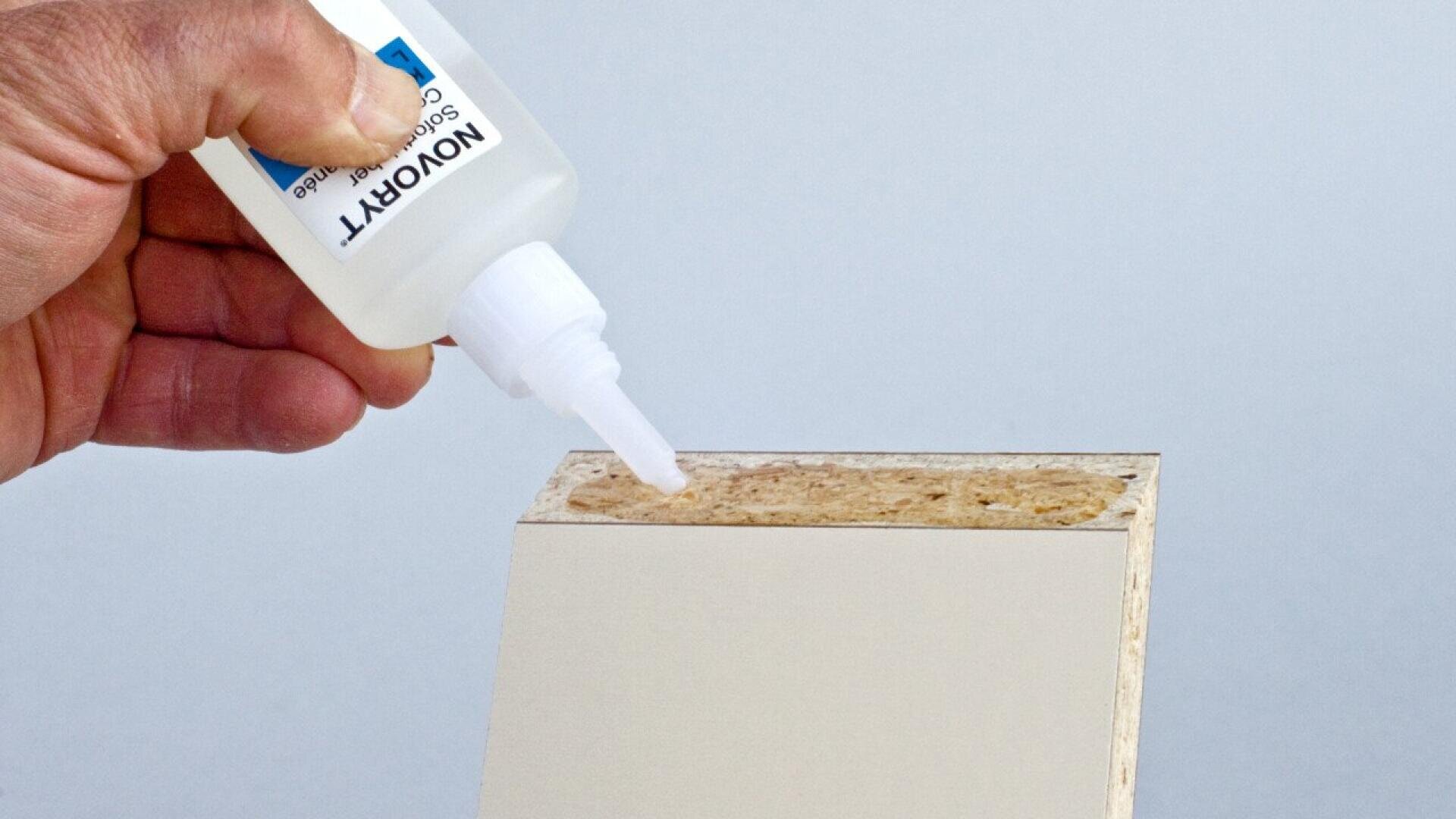 Une main applique de la colle à partir d'un flacon compressible sur le bord rugueux d'un panneau de particules avec une surface stratifiée blanche.