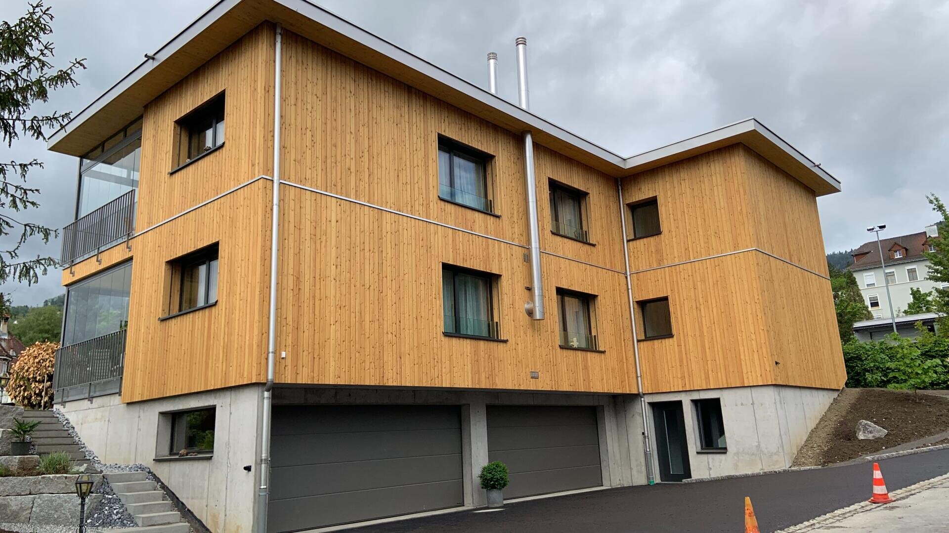 Ein modernes dreistöckiges Gebäude mit Holzvertäfelung und Betonsockel, mit drei Garagentoren und mehreren Fenstern, auf einer abschüssigen Zufahrt mit Grünpflanzen und einem orangefarbenen Verkehrskegel in der Nähe.