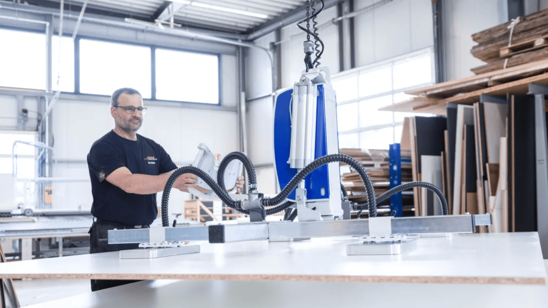 Ein Mann bedient einen großen Roboterarm mit Saugnäpfen, um eine weiße Platte in einer geräumigen, gut beleuchteten Werkstatt anzuheben, die mit Holzbearbeitungsgeräten und Holzstapeln gefüllt ist.
