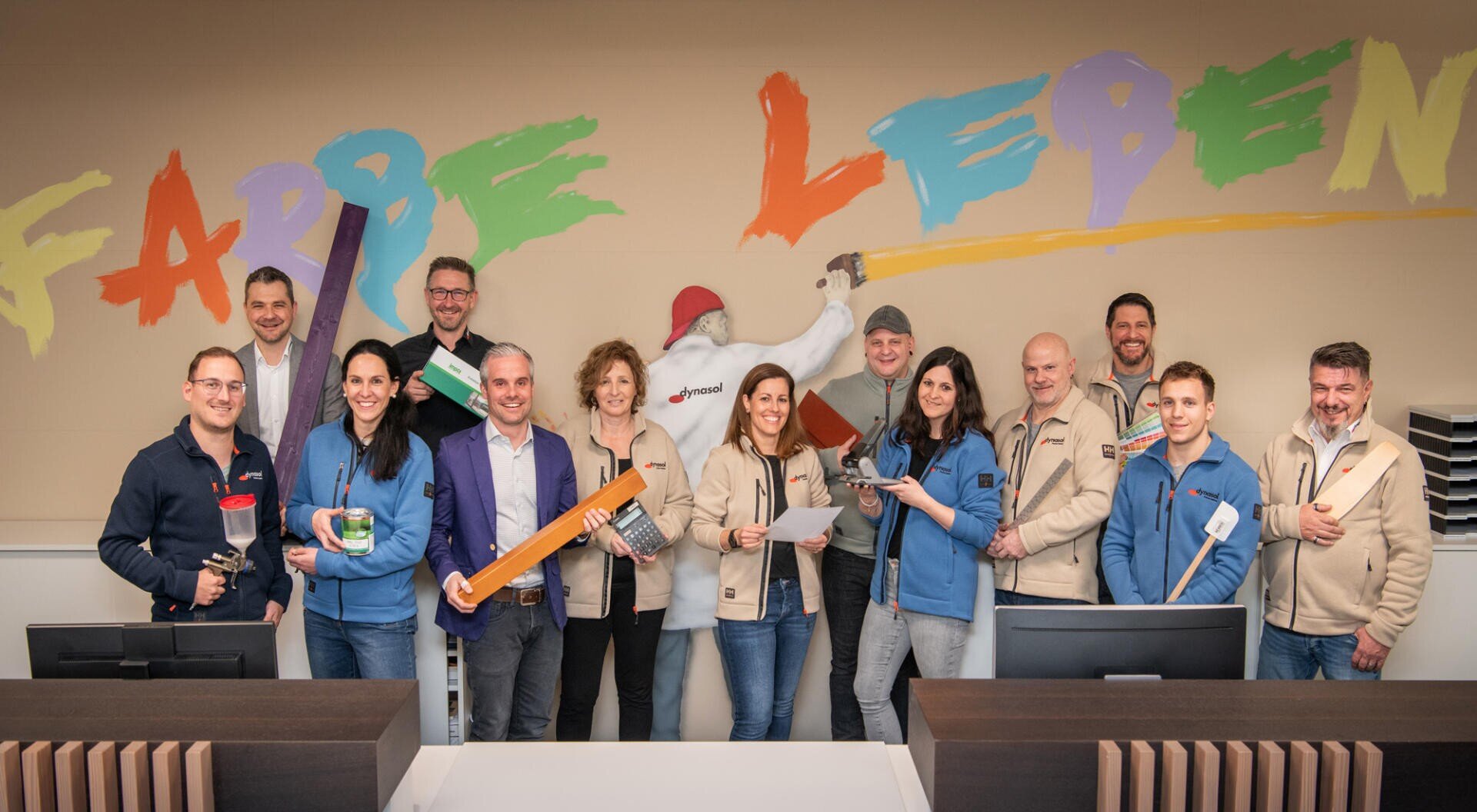 Un groupe de treize personnes souriantes se tient devant une fresque murale colorée portant l'inscription "VIE EN COULEUR". Ils tiennent des outils de peinture à la main et portent des vestes beiges ou bleues. Sur le mur derrière eux est peint un personnage de peintre.