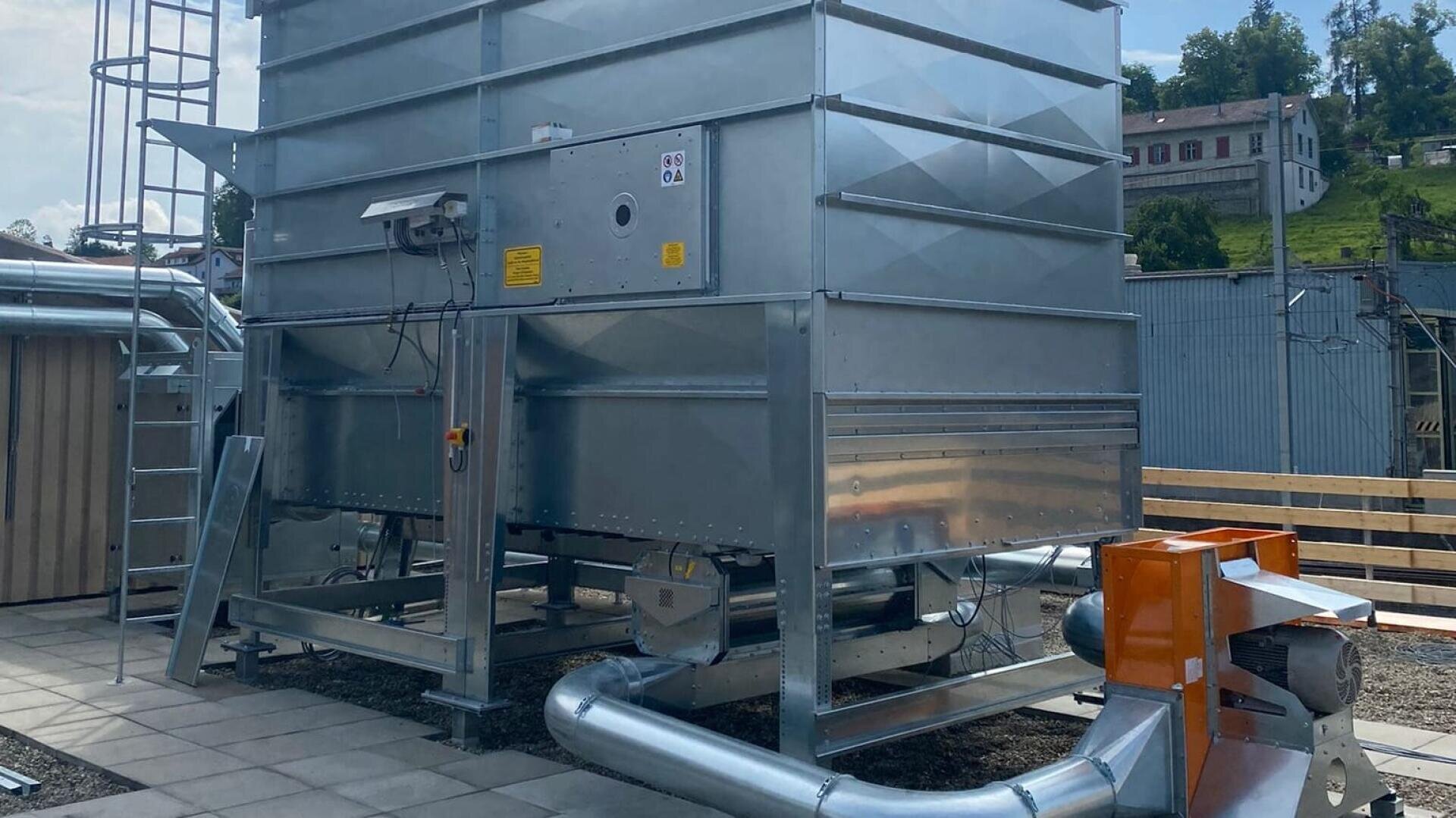 Große industrielle Metallstruktur mit Rohren und einem Ventilatorsystem im Freien, umgeben von Gebäuden und Grünanlagen. Eine Leiter und Warnschilder sind an der Anlage angebracht. Der Himmel ist klar und sonnig.