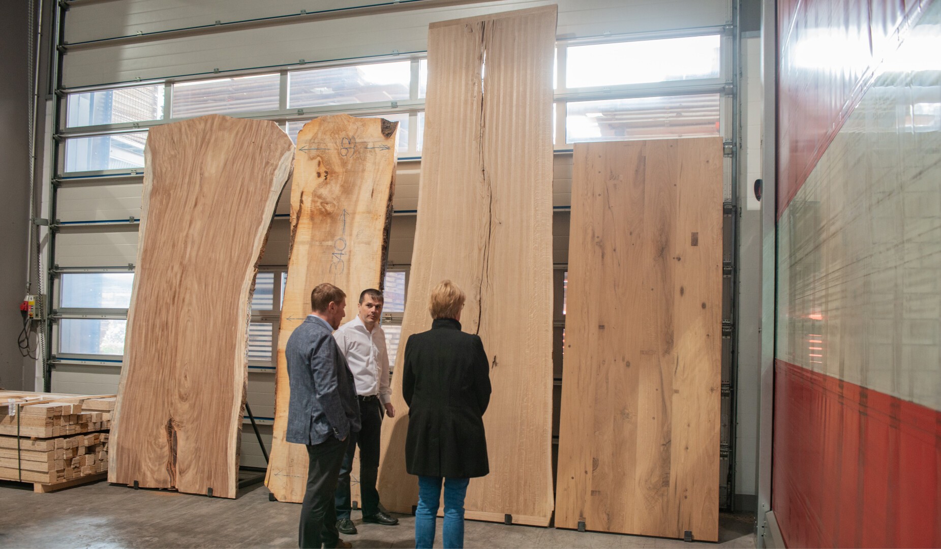 Drei Personen stehen im Innenbereich vor vier großen Holzplatten, die an einer Wand lehnen, diskutieren oder betrachten das Holz. Seitlich in der Werkstatt sind Stapel geschnittener Hölzer zu sehen.