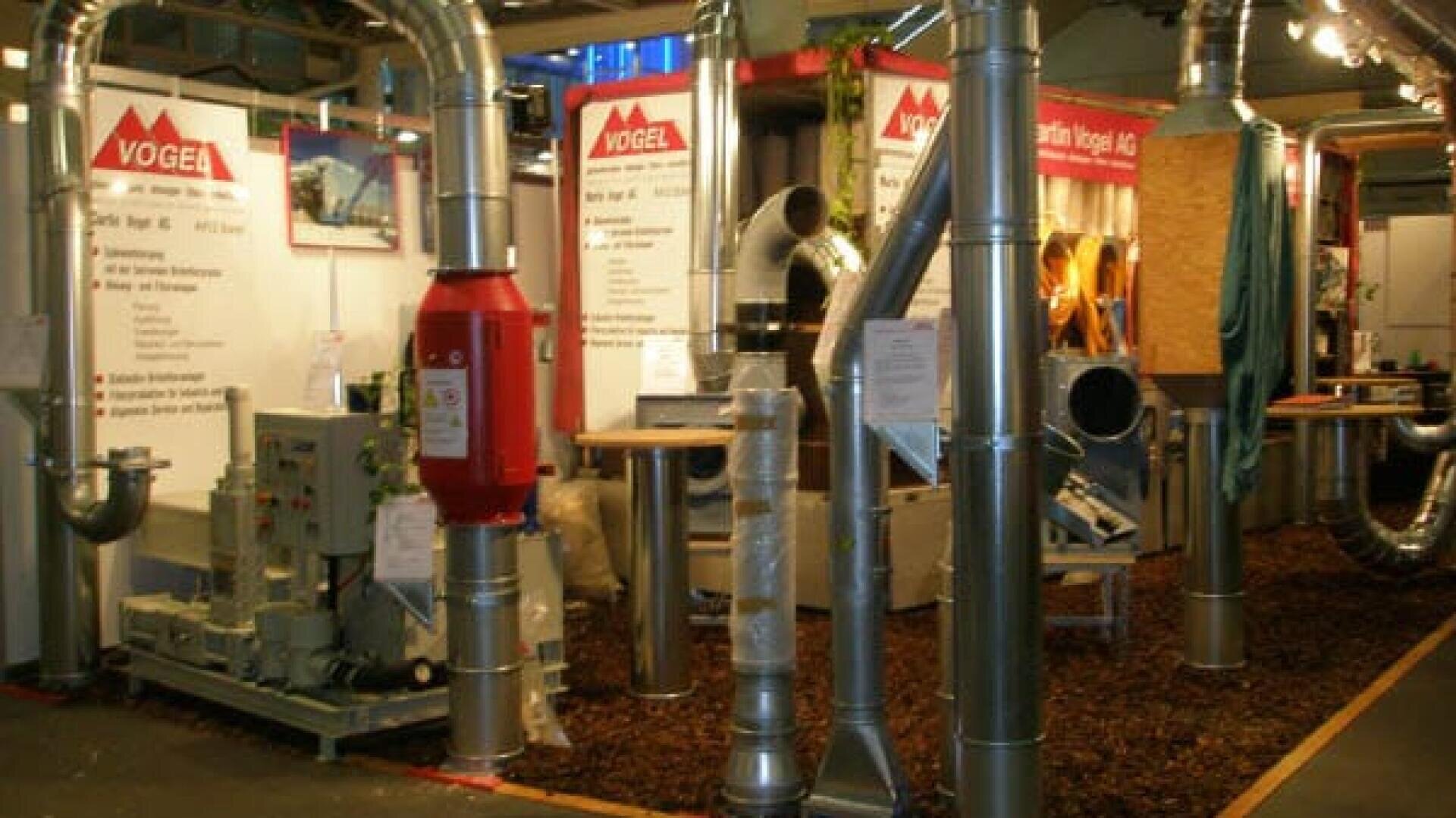 Industrieller Messestand mit verschiedenen Metallrohren, Maschinen und Lüftungsanlagen, im Hintergrund "VOGEL"-Banner und Informationsplakate. Der Boden ist mit Holzspänen bedeckt.