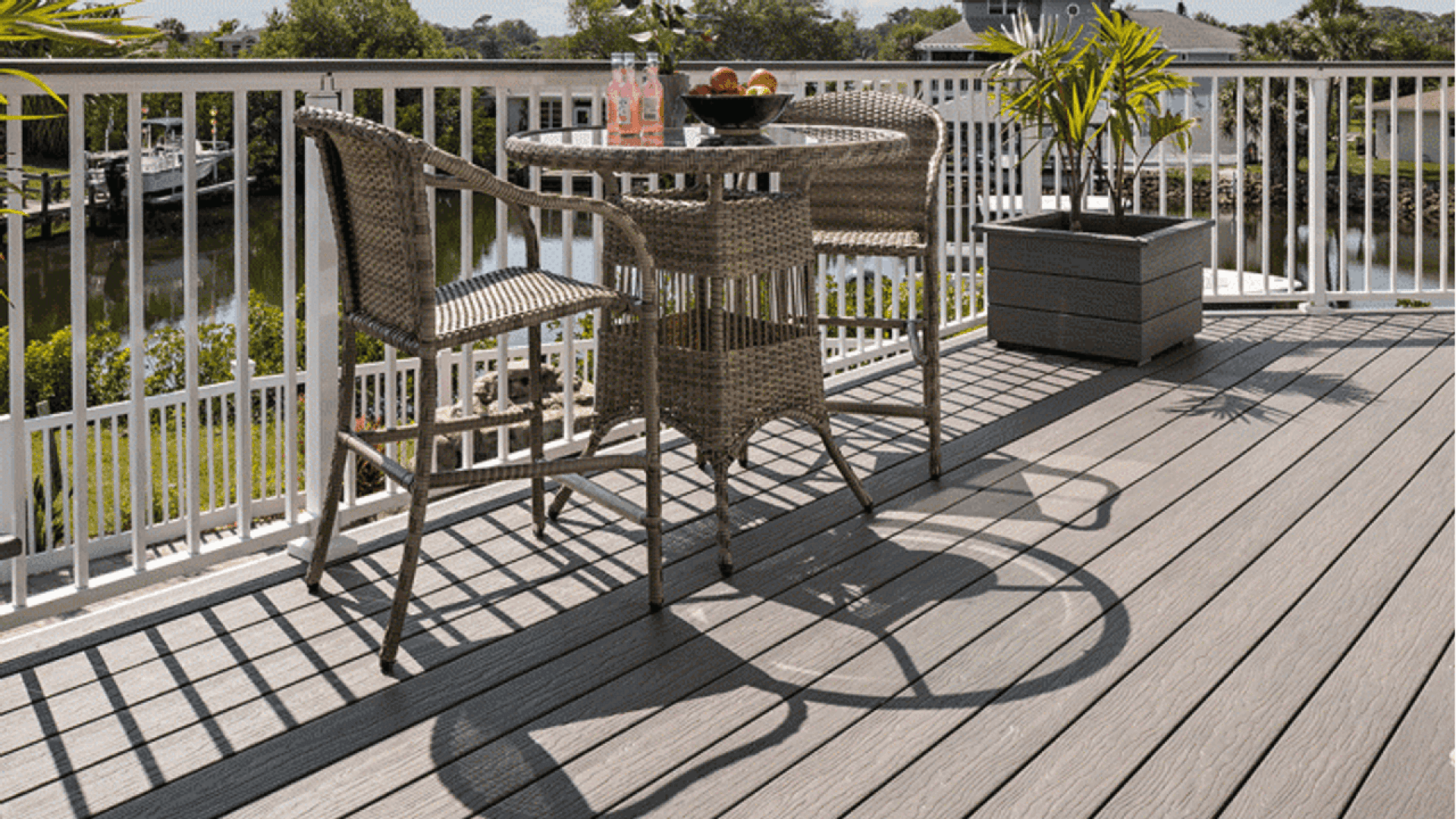 Une petite table d'extérieur avec deux chaises en osier est posée sur une terrasse, avec des boissons et un bol de fruits sur la table. Une plante en pot se trouve à proximité. La terrasse donne sur un plan d'eau et des arbres verts.