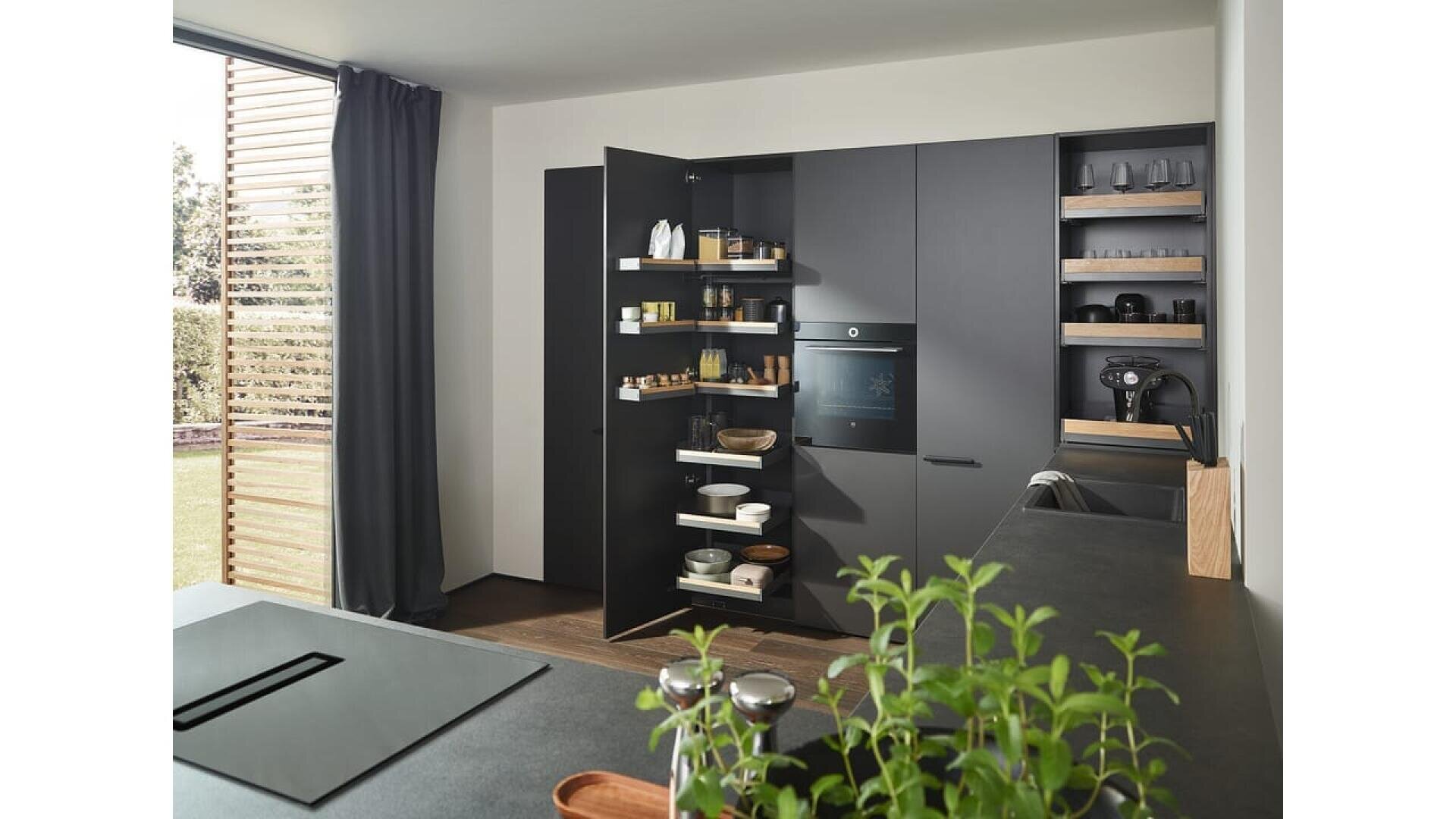 Cuisine moderne avec des armoires noires mates, un garde-manger ouvert rempli de plats et d'aliments, un four encastré, des appareils électroménagers sur le comptoir et une plante aromatique au premier plan. Une grande fenêtre laisse entrer la lumière naturelle.