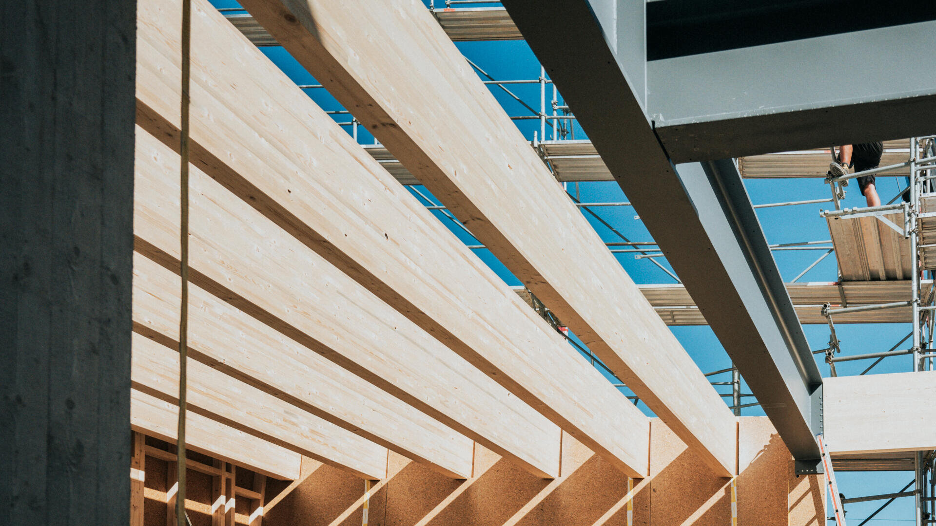 Holzbalken und Metallgerüste auf einer Baustelle unter strahlend blauem Himmel, mit geometrischen Linien und Schatten, die eine abstrakte architektonische Komposition ergeben.