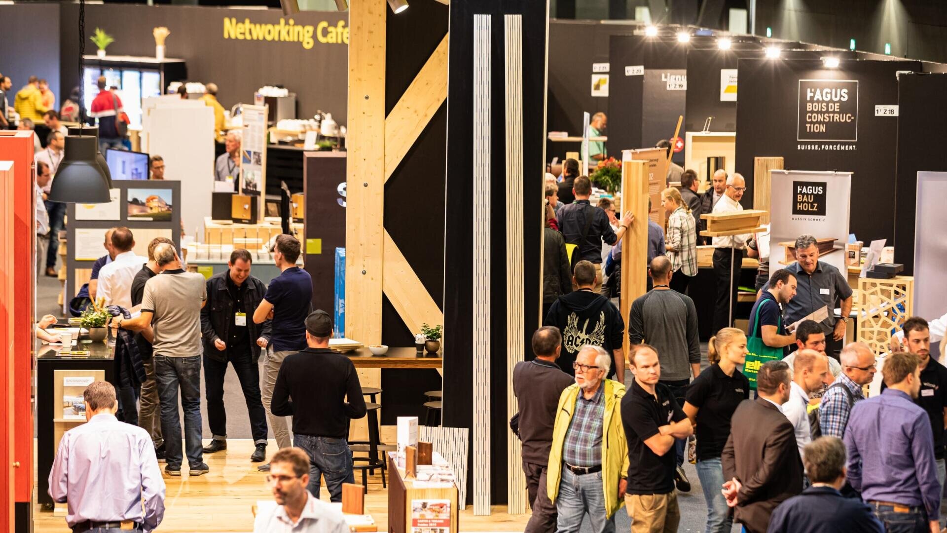 Un hall d'exposition très fréquenté où des groupes de personnes discutent et travaillent en réseau autour de divers stands et expositions, avec des bannières, des affiches et un panneau "Networking Café" visible en arrière-plan.