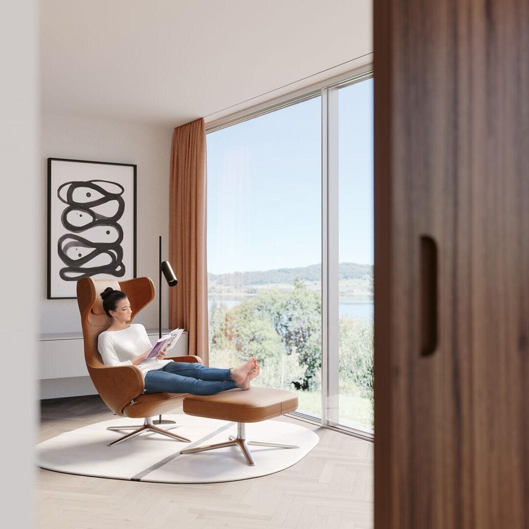 Une femme se prélasse sur une chaise brune avec un ottoman, lisant un livre dans une pièce moderne et lumineuse avec de grandes fenêtres, des rideaux orange, un décor minimaliste et une vue pittoresque sur l'extérieur.