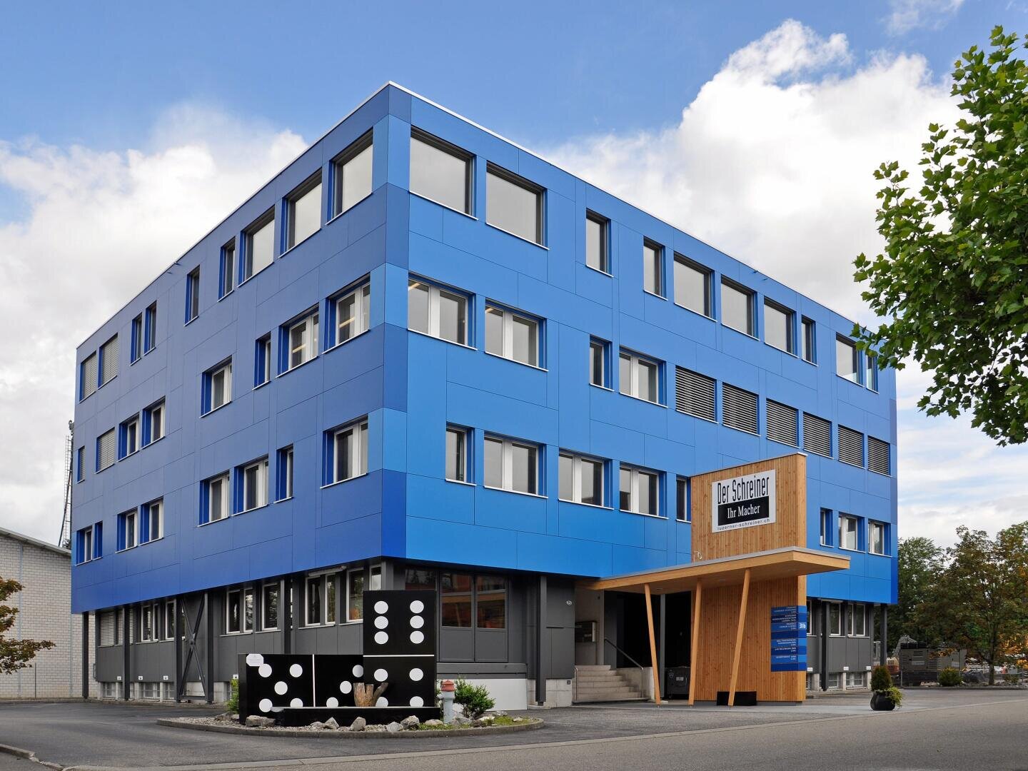 Ein modernes, vierstöckiges Gebäude mit hellblauer Vertäfelung und vielen Fenstern. Der Eingang hat hölzerne Akzente und ein Schild mit der Aufschrift Die Echten über der Tür. Eine große Dominoskulptur ziert die Fassade. Bäume und ein bewölkter Himmel umgeben das Gebäude.