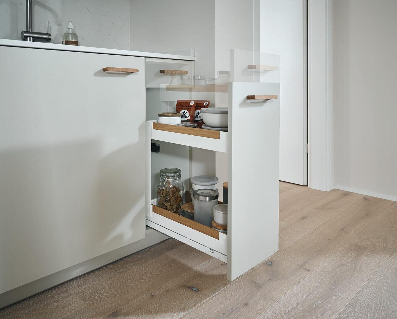 Ein moderner weißer Küchenschrank mit einer schmalen ausziehbaren Schublade, die Gläser, Behälter und Gewürze enthält, vor einem hellen Holzboden.