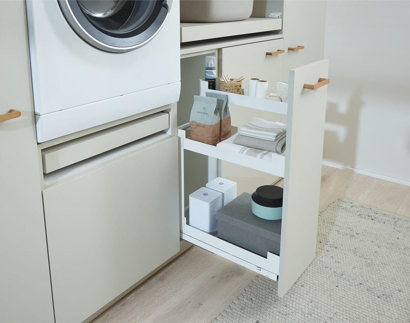 Ein ausziehbares Regal neben der Waschmaschine bietet Platz für Wäsche, Handtücher und Kartons in einer modernen Waschküche mit hellen Schränken und Holzboden.