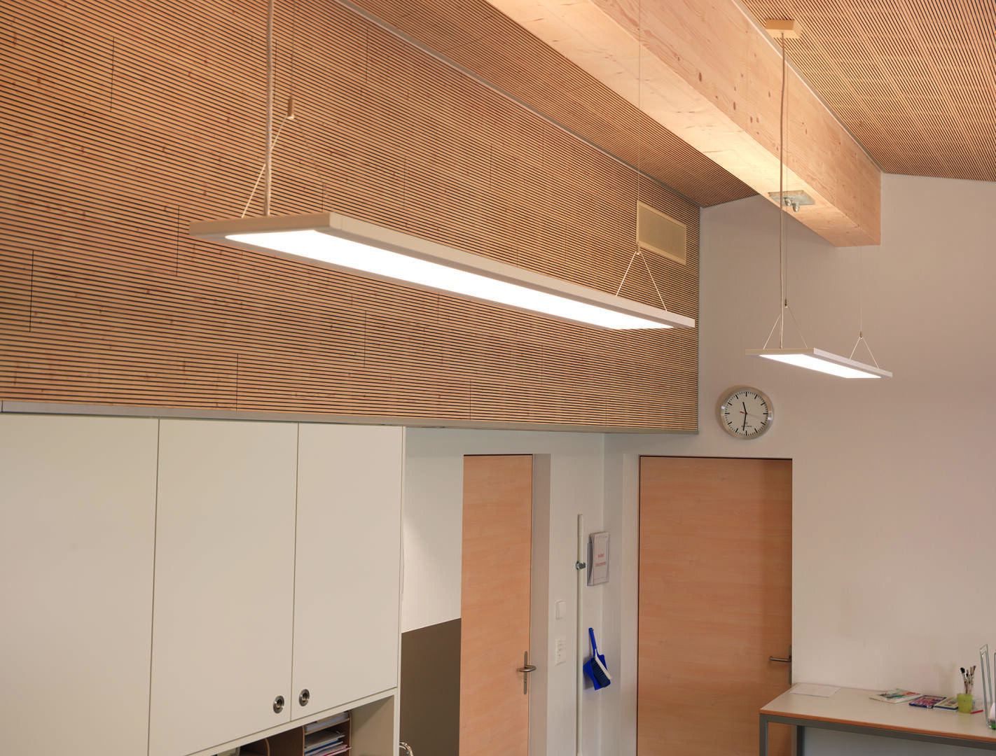 Moderne Büroeinrichtung mit Holztüren, weißen Schränken, einer Wanduhr und zwei rechteckigen Deckenleuchten. Die Decke und die oberen Wände sind mit horizontalen Holzlatten versehen, die zu einem minimalistischen Design beitragen.