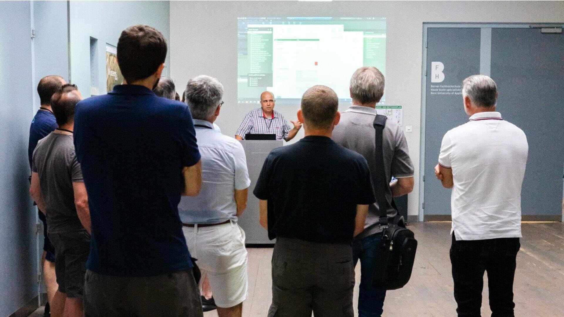 Un groupe de personnes se tient debout et écoute un orateur faire une présentation dans une salle. L'orateur se trouve sur un podium et derrière lui se trouve un écran projeté qui affiche un logiciel ou un site web.